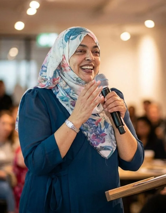Nurjahan Khatun, coach & speaker. wearing a floral hijab and blue dress speaking into a microphone at an indoor event with a blurred audience in the background.