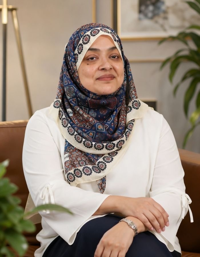 Nurjahan Khatun, coach & speaker. wearing a colourful patterned hijab and a white blouse, sitting on a chair in a well-lit room with a plant and artwork in the background.