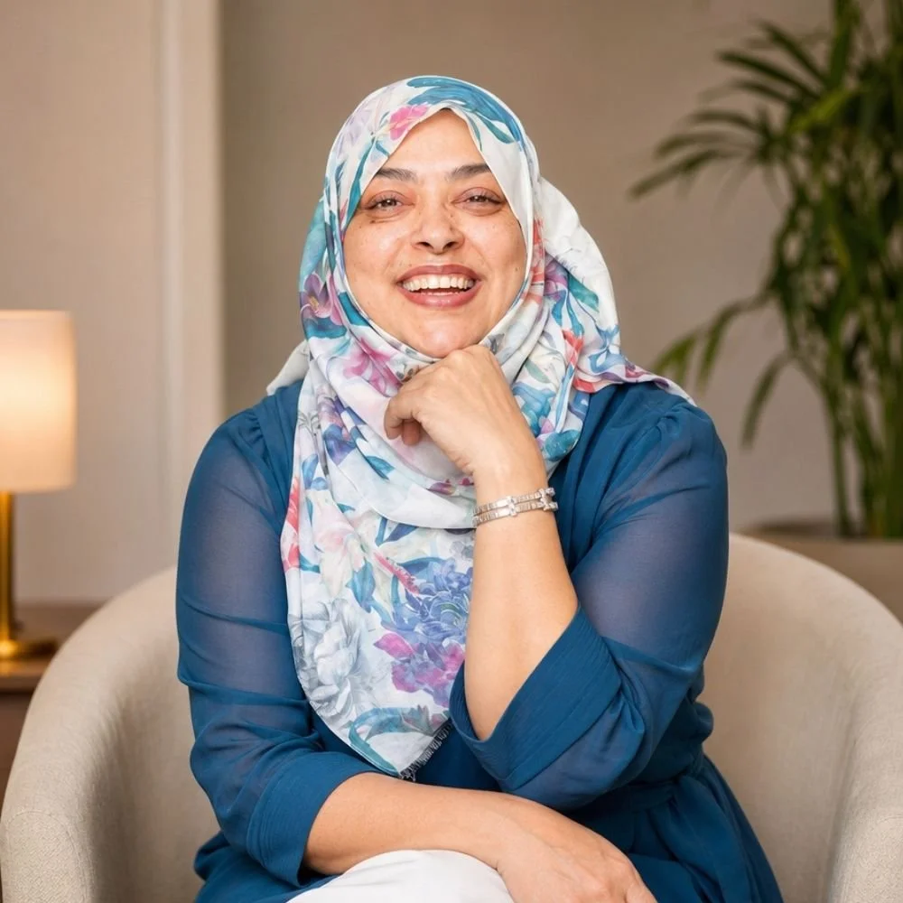 Nurjahan Khatun, coach & speaker. wearing a floral headscarf and a blue dress, sitting on a beige chair, with a warm indoor setting featuring a lamp and a green plant in the background.