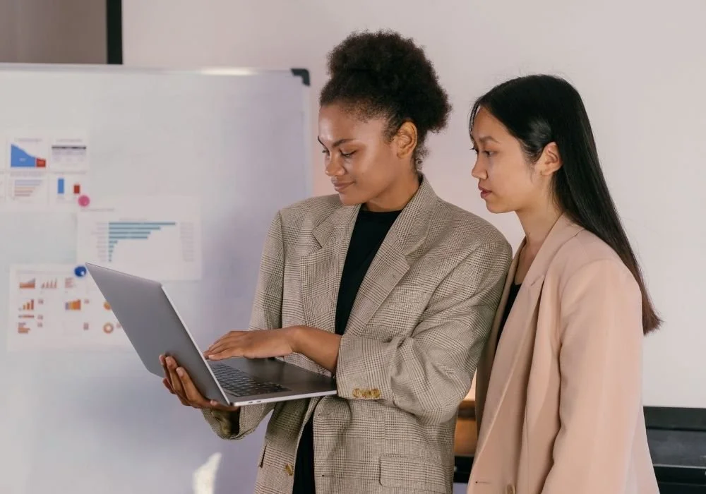 Two women in business attire standing close together, looking at a laptop screen. One woman with curly hair is holding the laptop, and the other woman with straight hair is looking at the screen. In the background, there is a whiteboard with charts and graphs.