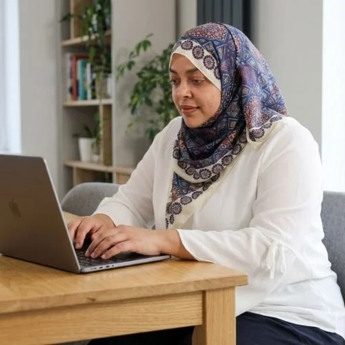 Nurjahan Khatun, coach & speaker. wearing a patterned headscarf working on a laptop at a wooden table in a home office.
