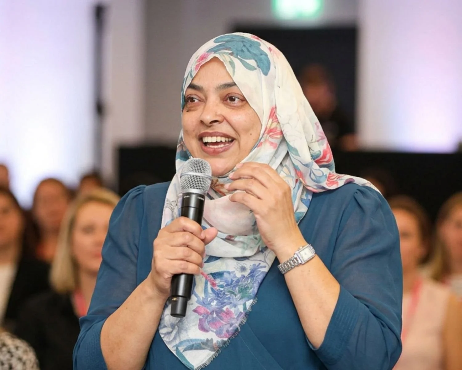 Nurjahan Khatun, coach & speaker. wearing a floral headscarf and blue top speaking into a microphone during an indoor event.