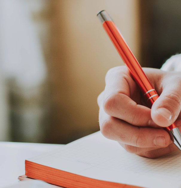Close-up of a hand holding a red pen writing in an open notebook with a white page and red edges.