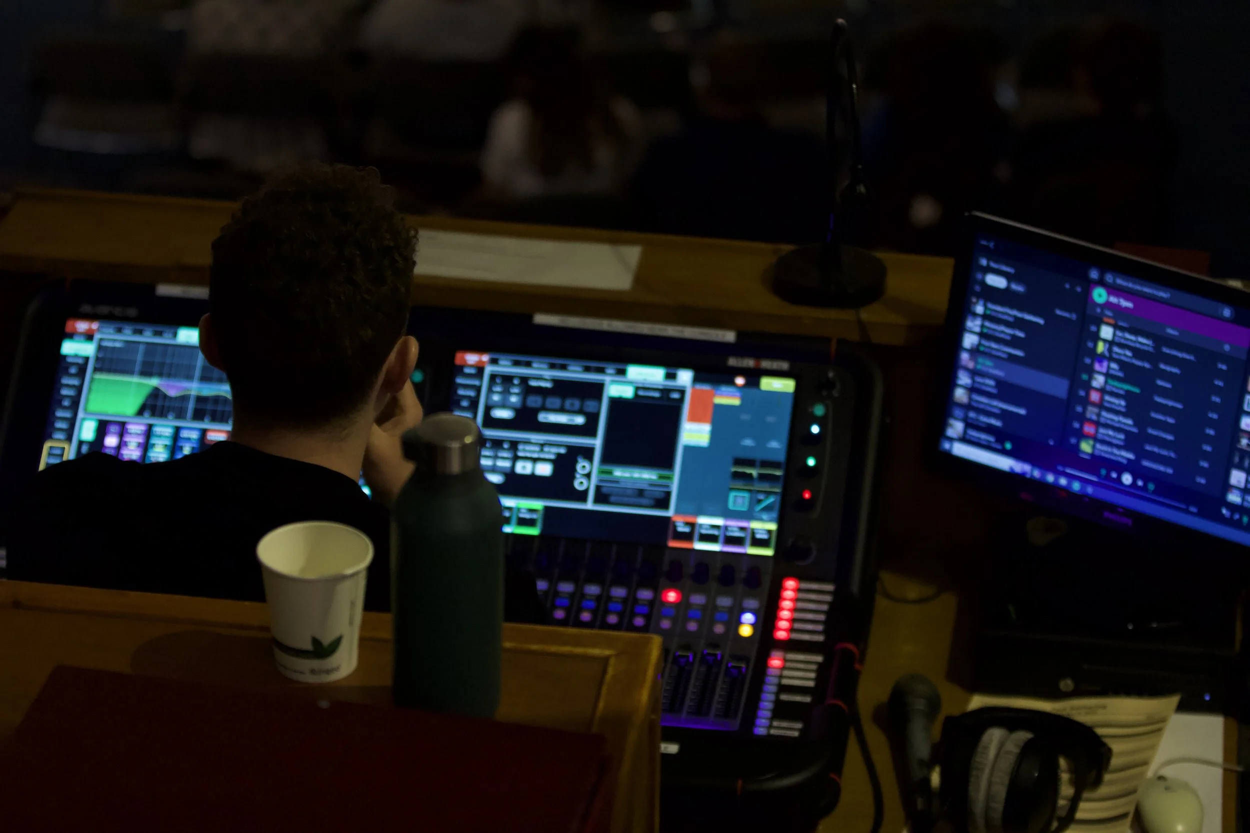 A person operating sound equipment at a live event, with multiple screens showing digital audio software, a microphone, a water bottle, and a paper cup on the table, in a dark venue setting.
