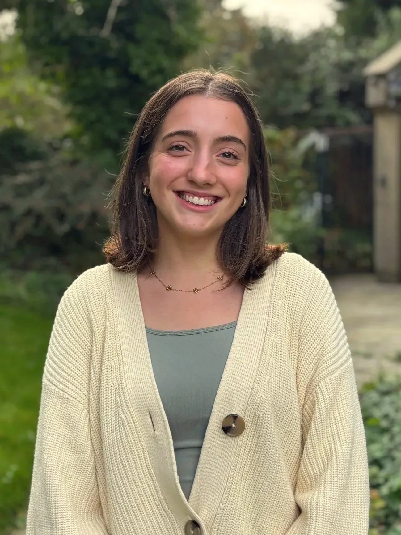A young woman with shoulder-length brown hair smiling outdoors in a garden during daytime, wearing a beige cardigan over a green top and a necklace with small flower charms.