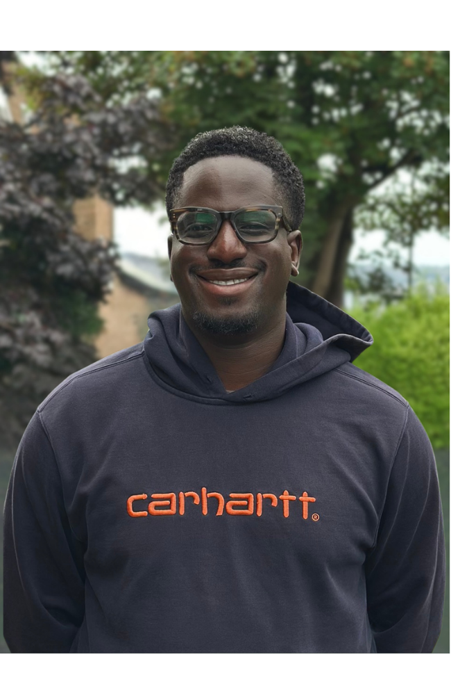 A smiling man with glasses and short curly hair, wearing a cream-colored sweatshirt with a graphic on the front, standing outdoors with greenery in the background.