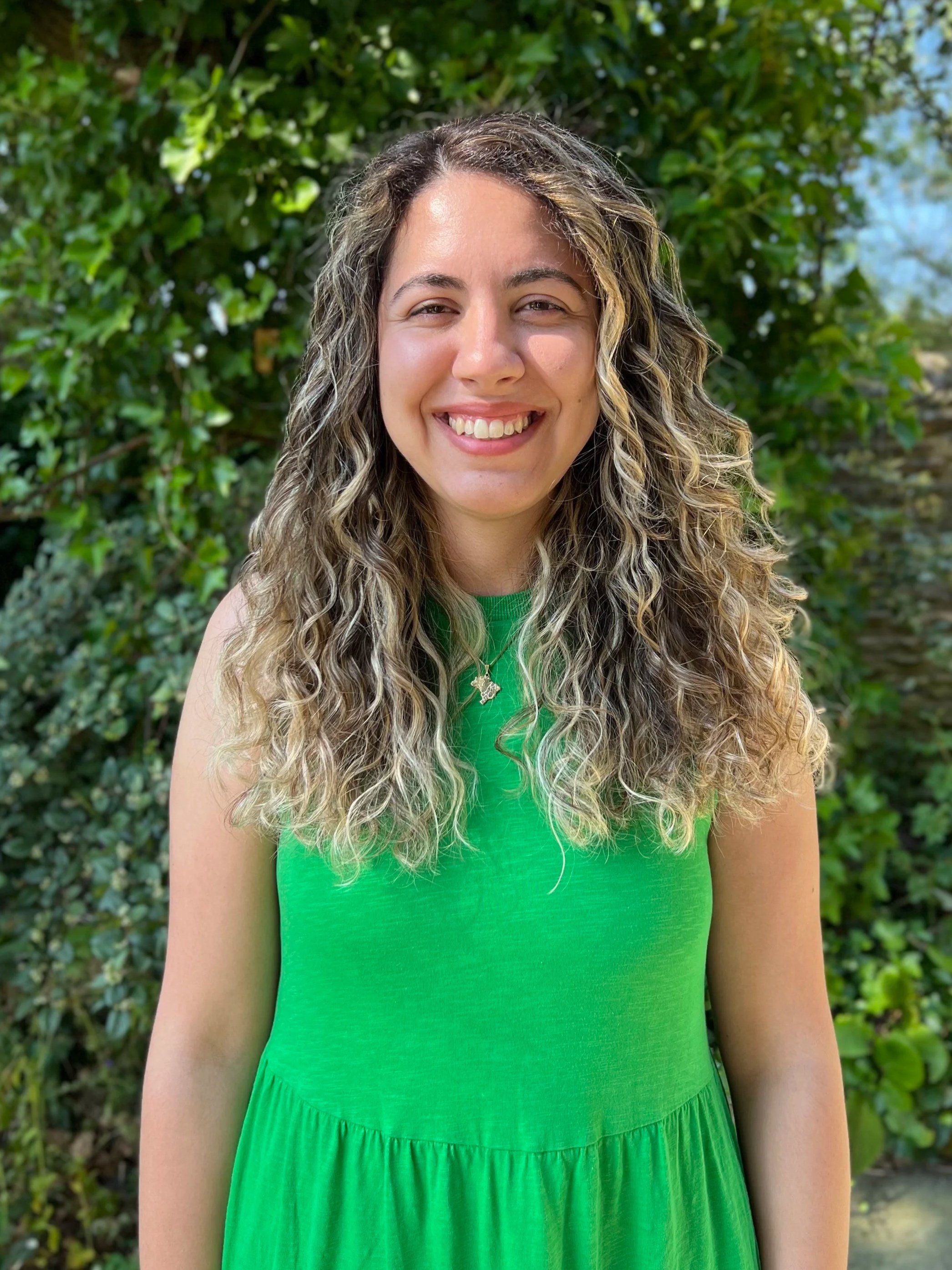 A woman with long, curly hair, wearing a green dress and a necklace with a small dog pendant, smiling outdoors against leafy green background.