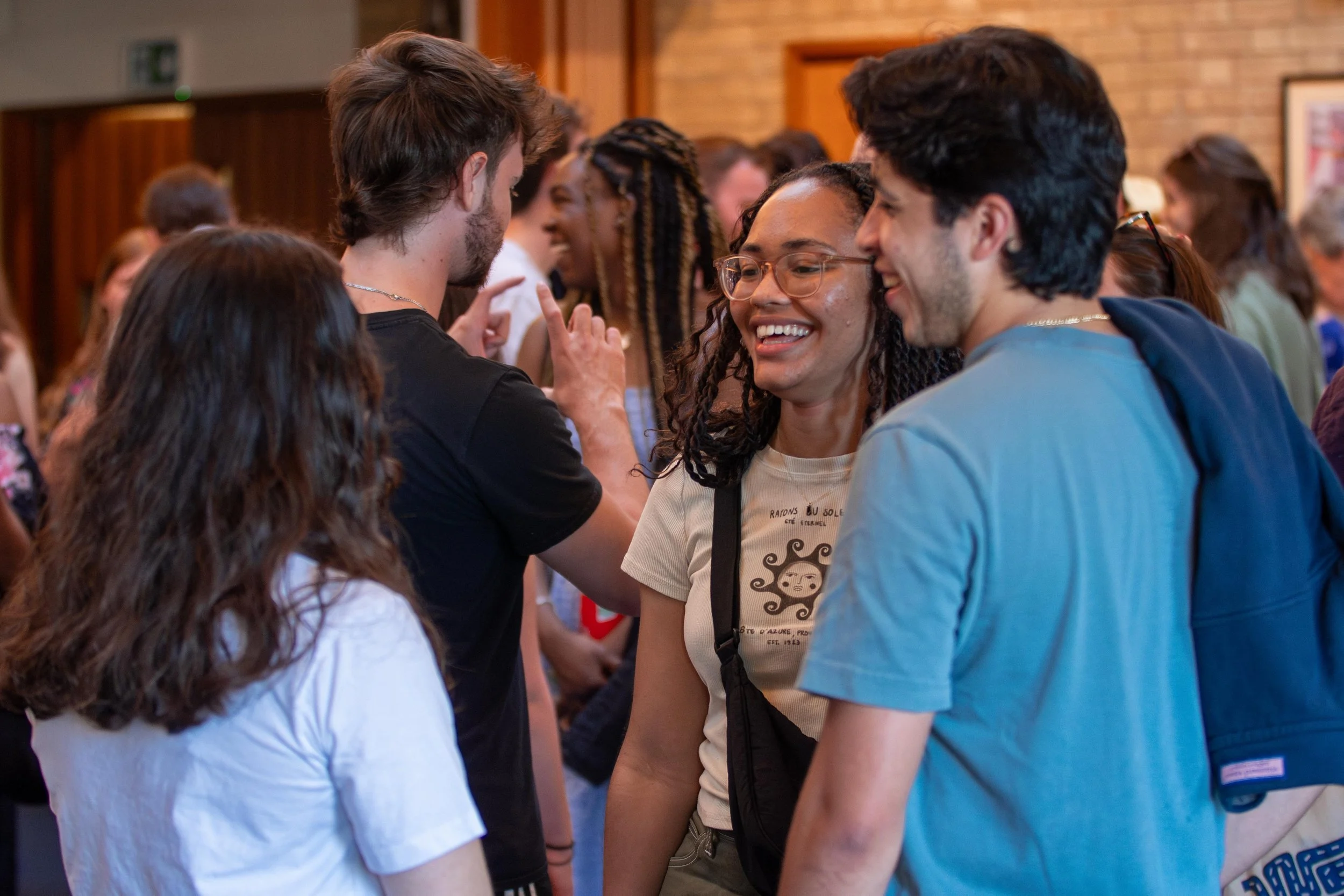 People smiling and talking closely in a group in an indoor setting, with wooden walls in the background.