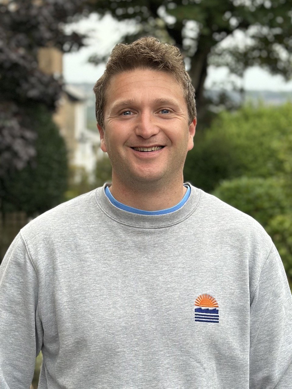 A smiling man wearing a gray sweatshirt with a small embroidered sunset and ocean waves design standing outdoors with trees and houses in the background.