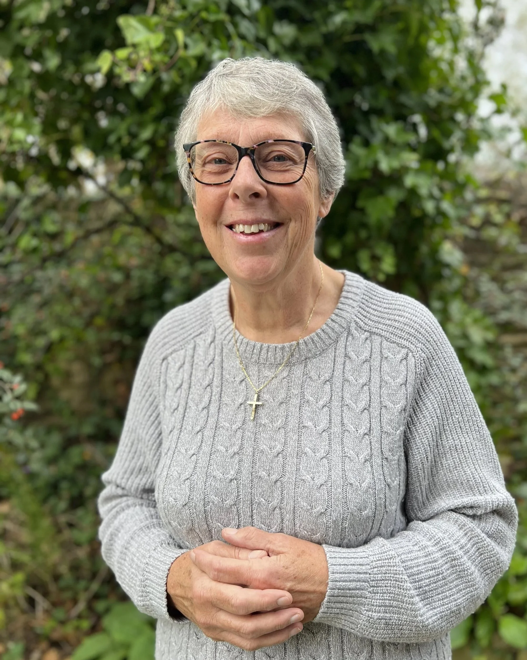 Older woman with short gray hair, glasses, and a cross necklace, smiling, wearing a gray sweater, standing outdoors in front of green foliage.