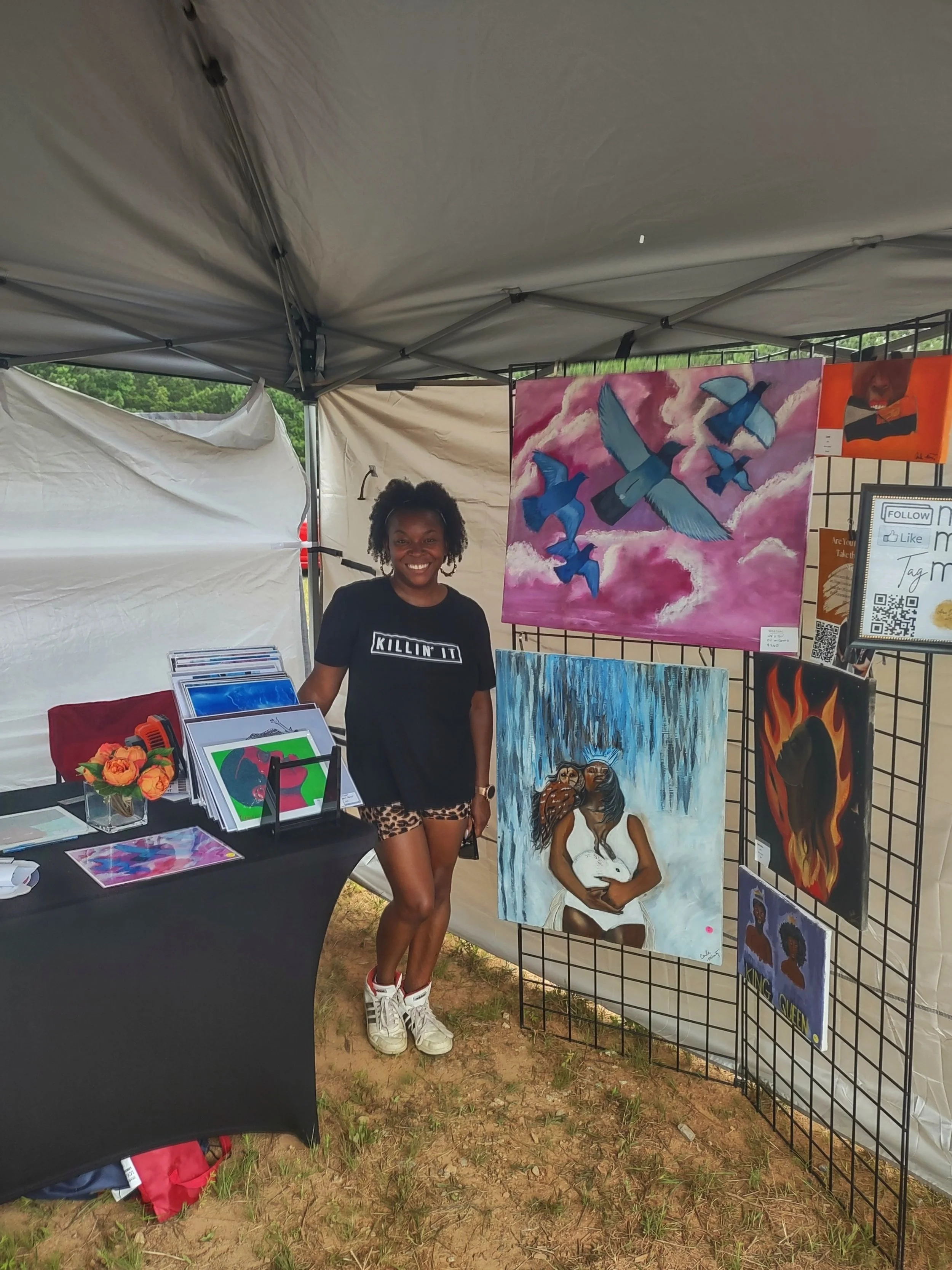 An artist standing in her booth with her oil paintings at an outdoor market