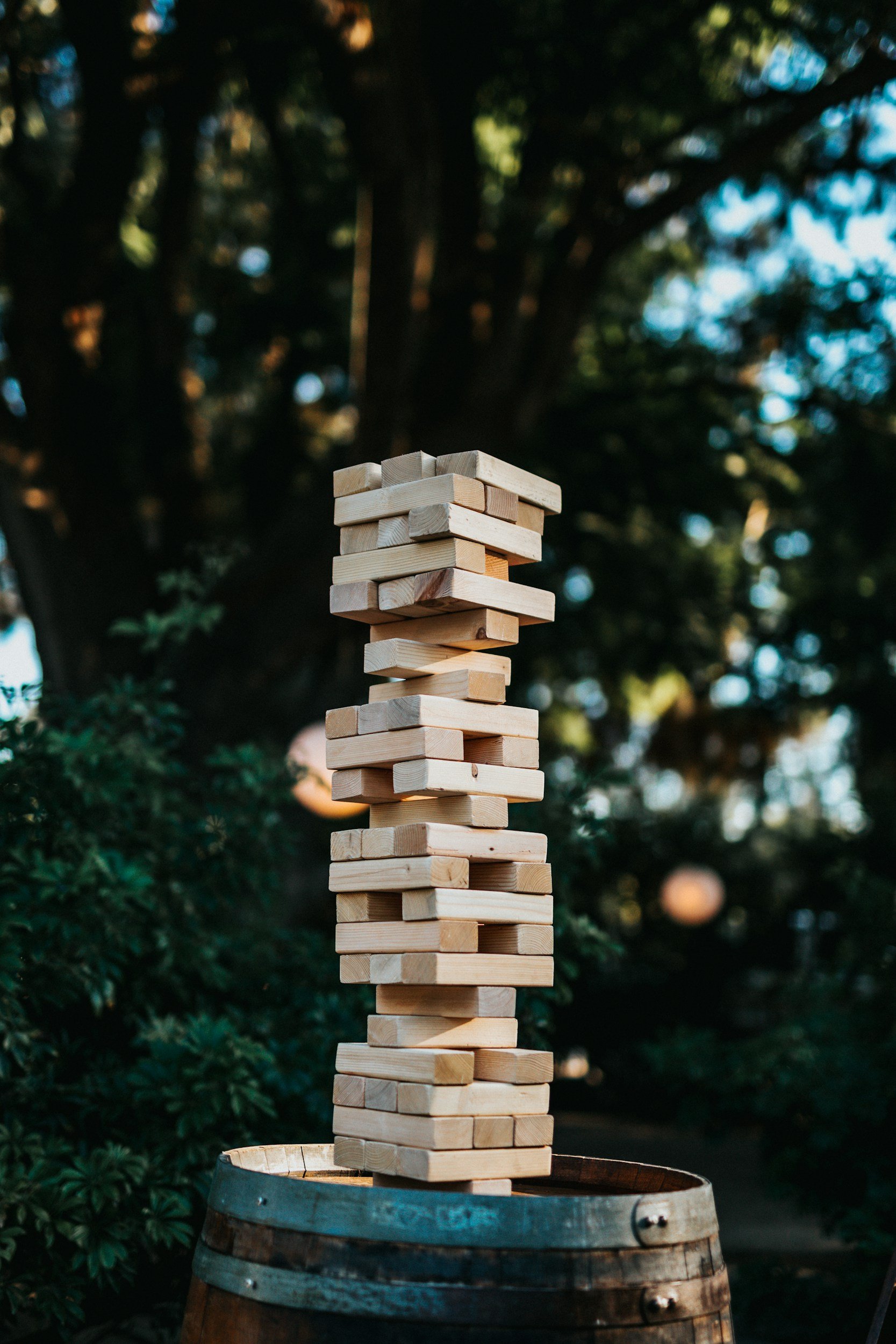A tower of wooden blocks stacked in a Jenga-like structure on top of a barrel outdoors with trees in the background.