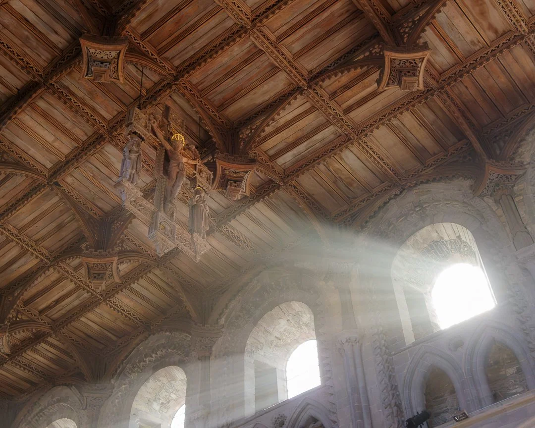 Sunlight streaming through arched windows into a grand interior with ornate woodwork and carved statues on the ceiling.