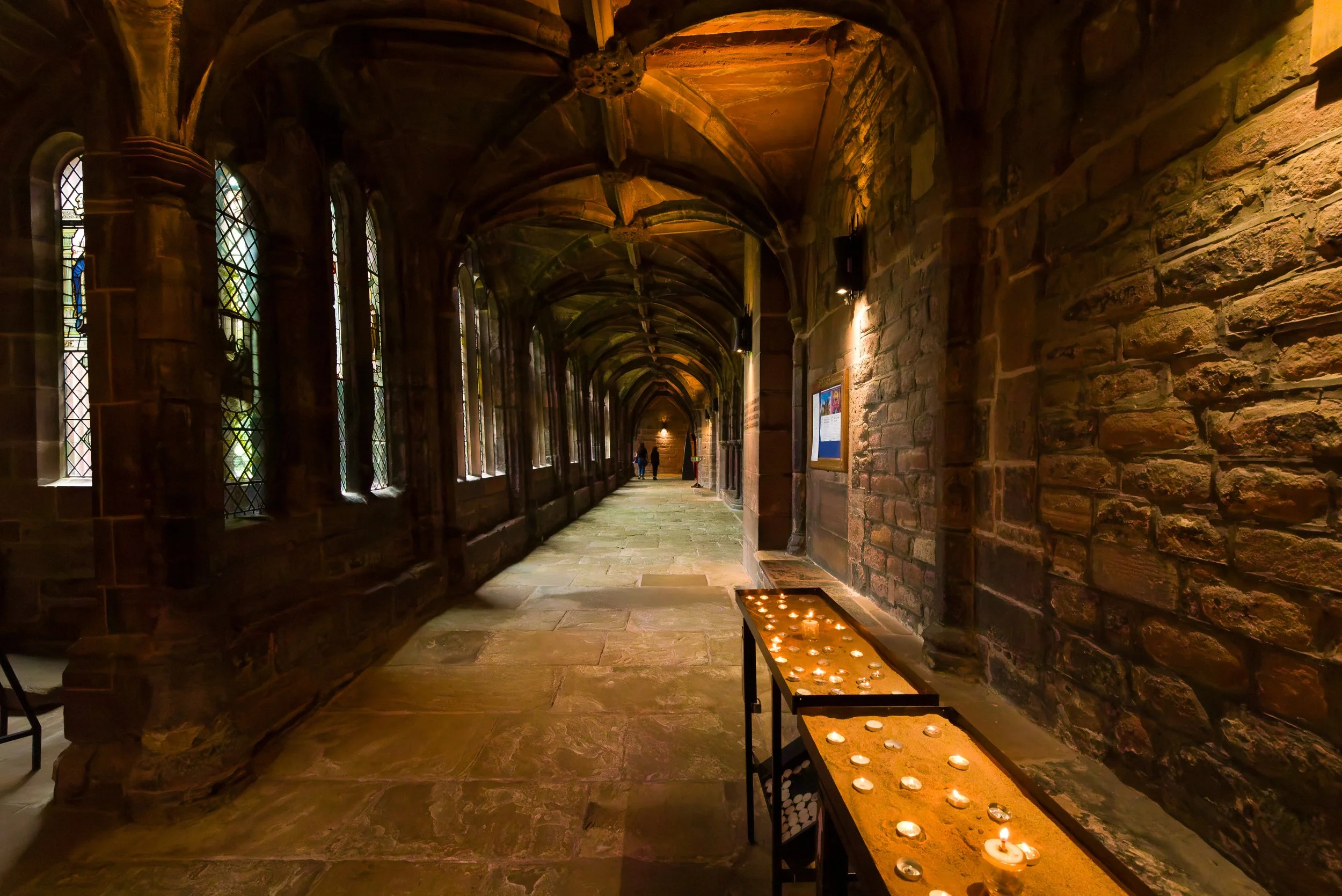 A dimly lit stone corridor with arched ceiling and stained glass windows on the left, with candles on a table on the right, and a few people walking in the distance.
