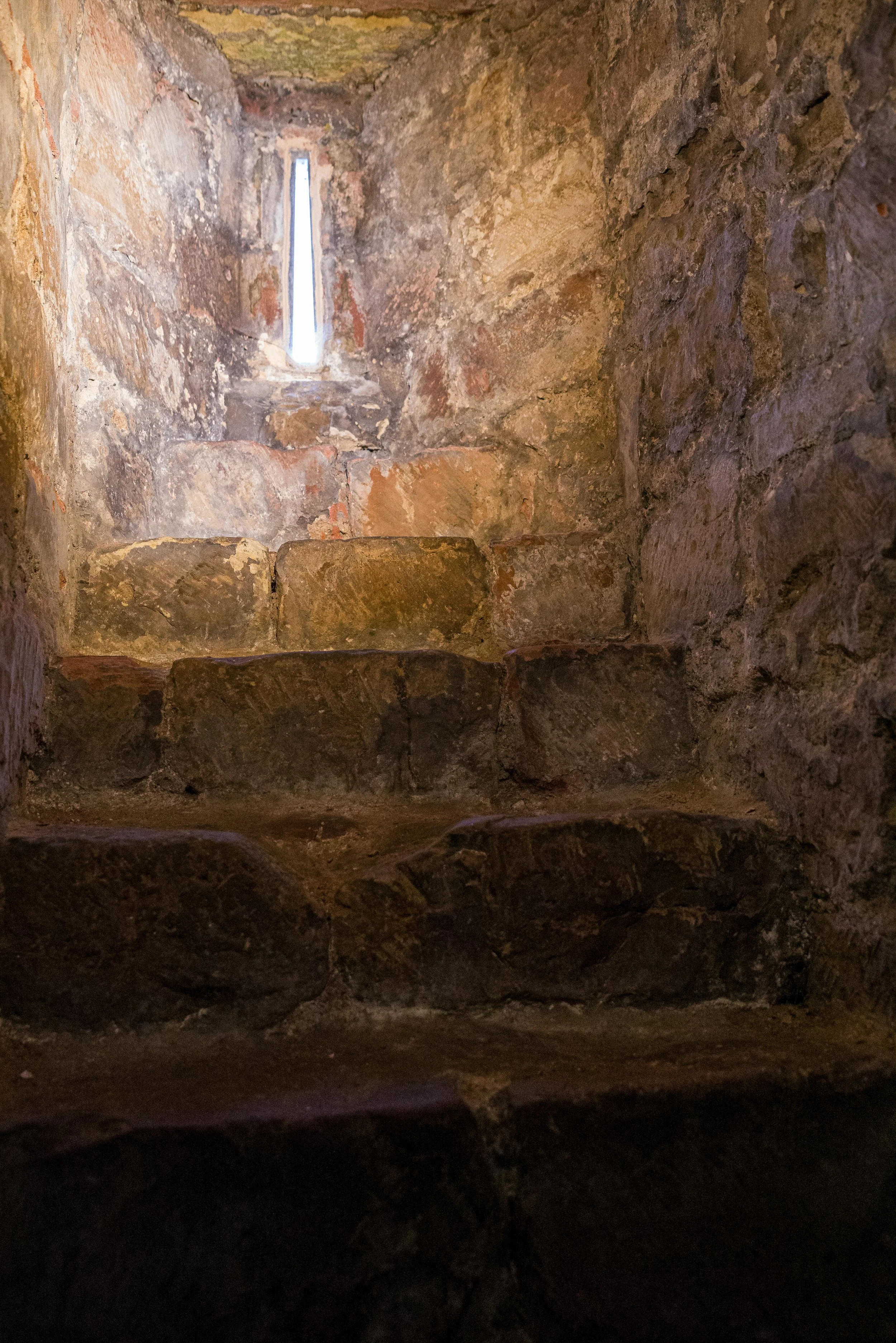 Stone staircase inside a small, enclosed stone chamber with a narrow vertical window at the top allowing light to enter.