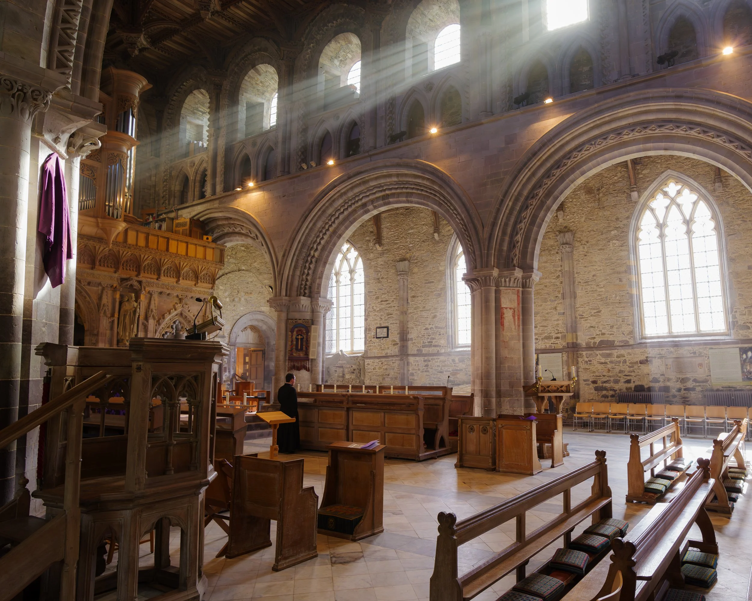 Interior of a historic church with high arched windows, stone walls, wooden pews, and sunlight streaming through the windows.