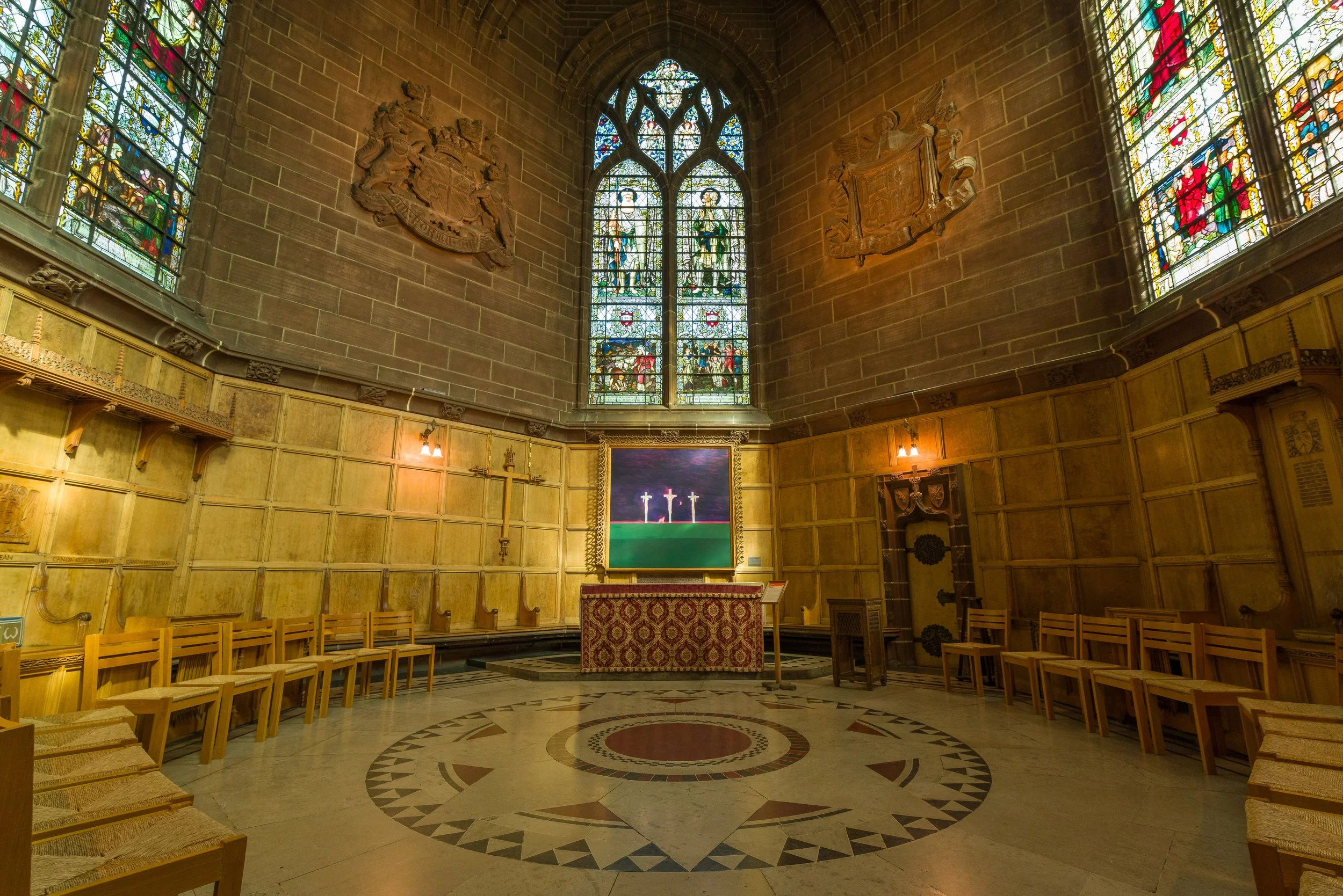 Interior of a church with stained glass windows, wooden wall paneling, a central altar with a painting, and chairs arranged along the walls.