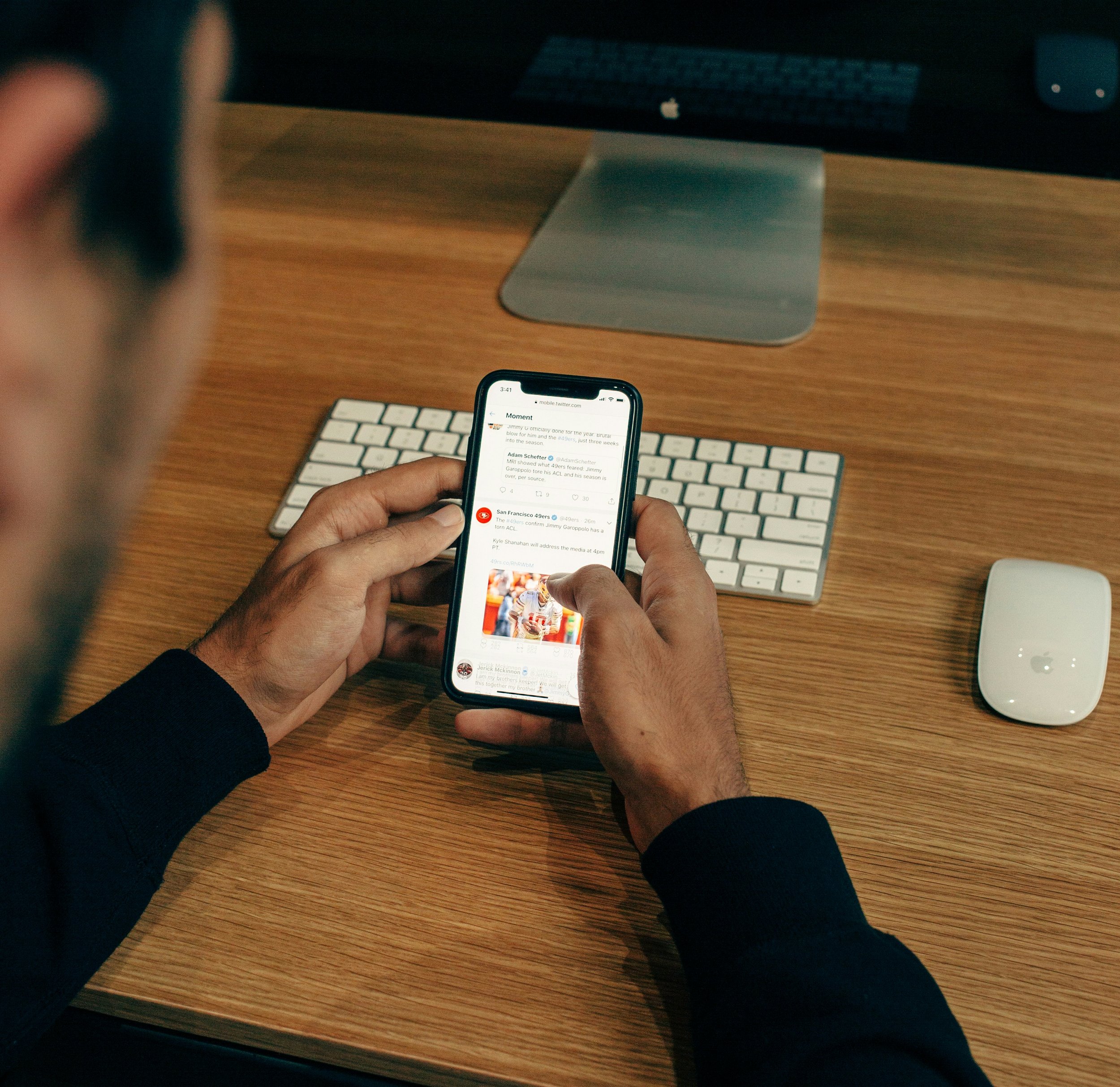 Person holding a smartphone while sitting at a desk with an iMac computer, keyboard, and Magic Mouse.