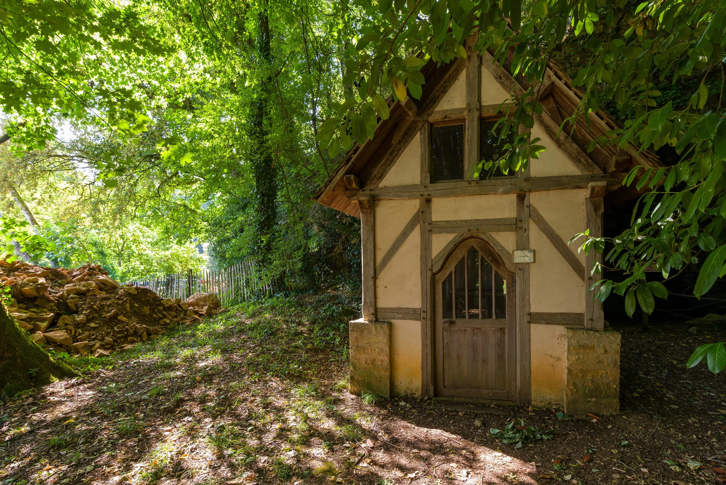 A small, rustic miniature house with a wooden door and windows, surrounded by lush greenery and trees, casting dappled sunlight on the forest floor.