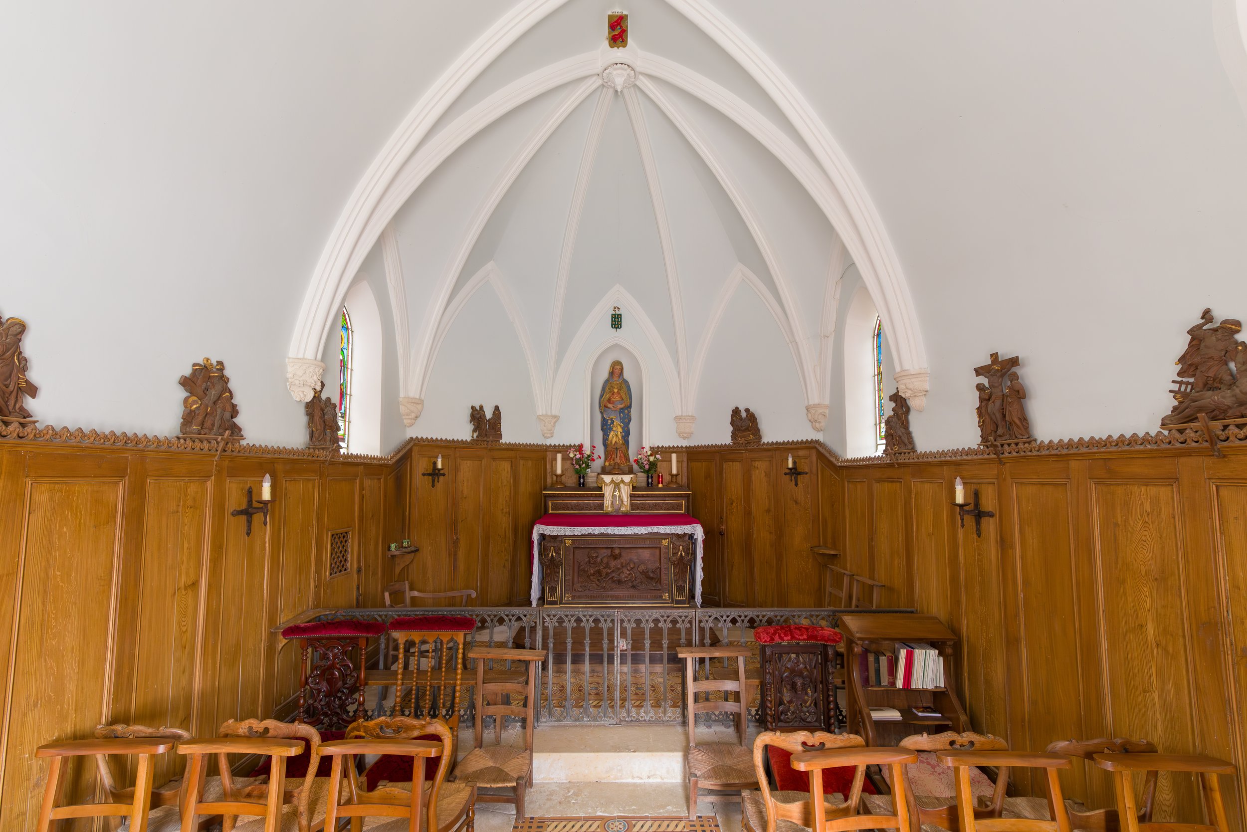 Interior of a small church or chapel with wooden pews and an altar featuring a statue of Mary in the center. The altar is decorated with flowers and candles. The ceiling has white gothic arches, adorned with wooden religious carvings along the walls 