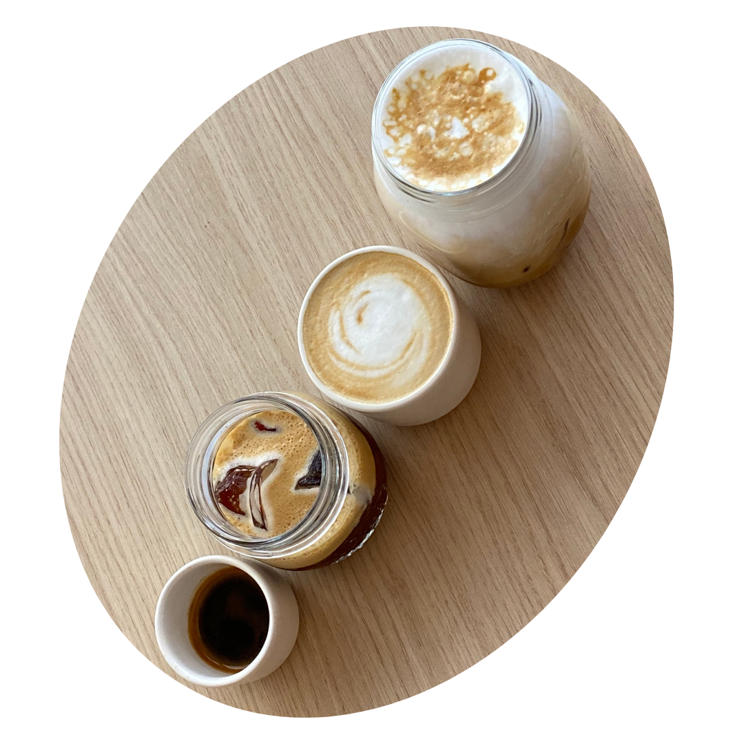 Cinq boissons chaudes différentes sur une table en bois, comprenant un espresso, un cappuccino, un café glacé, un latte macchiato, et un autre café dans un bocal.