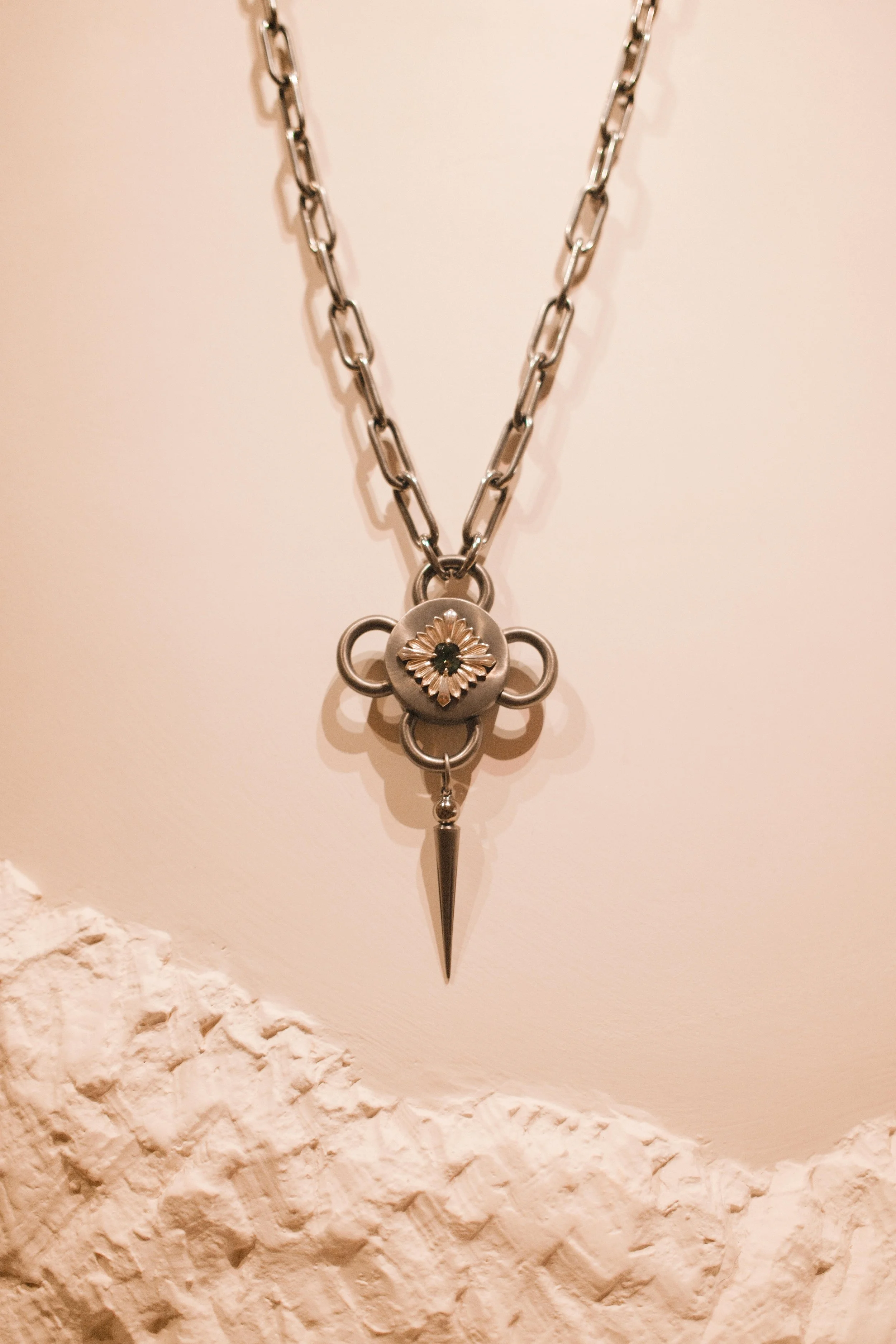 Silver necklace with a pendant featuring a floral design and a pointed spike hanging below, on a white textured background