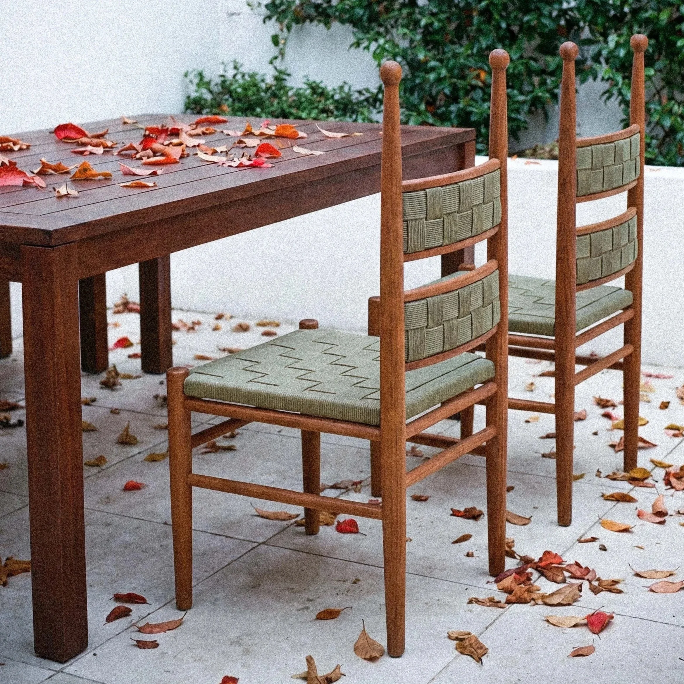 A wooden table with scattered autumn leaves, alongside three matching chairs with woven green backrests, on a tiled floor with more fallen leaves, outdoors near some green bushes.