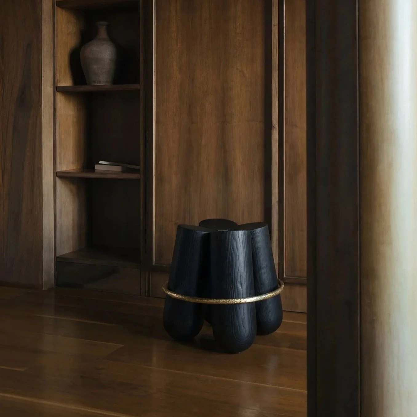 A dark wooden cabinet with open shelves, surrounding a closed door, and a black, modern stool with a gold ring in the foreground, on a wooden floor near a light-colored curtain.