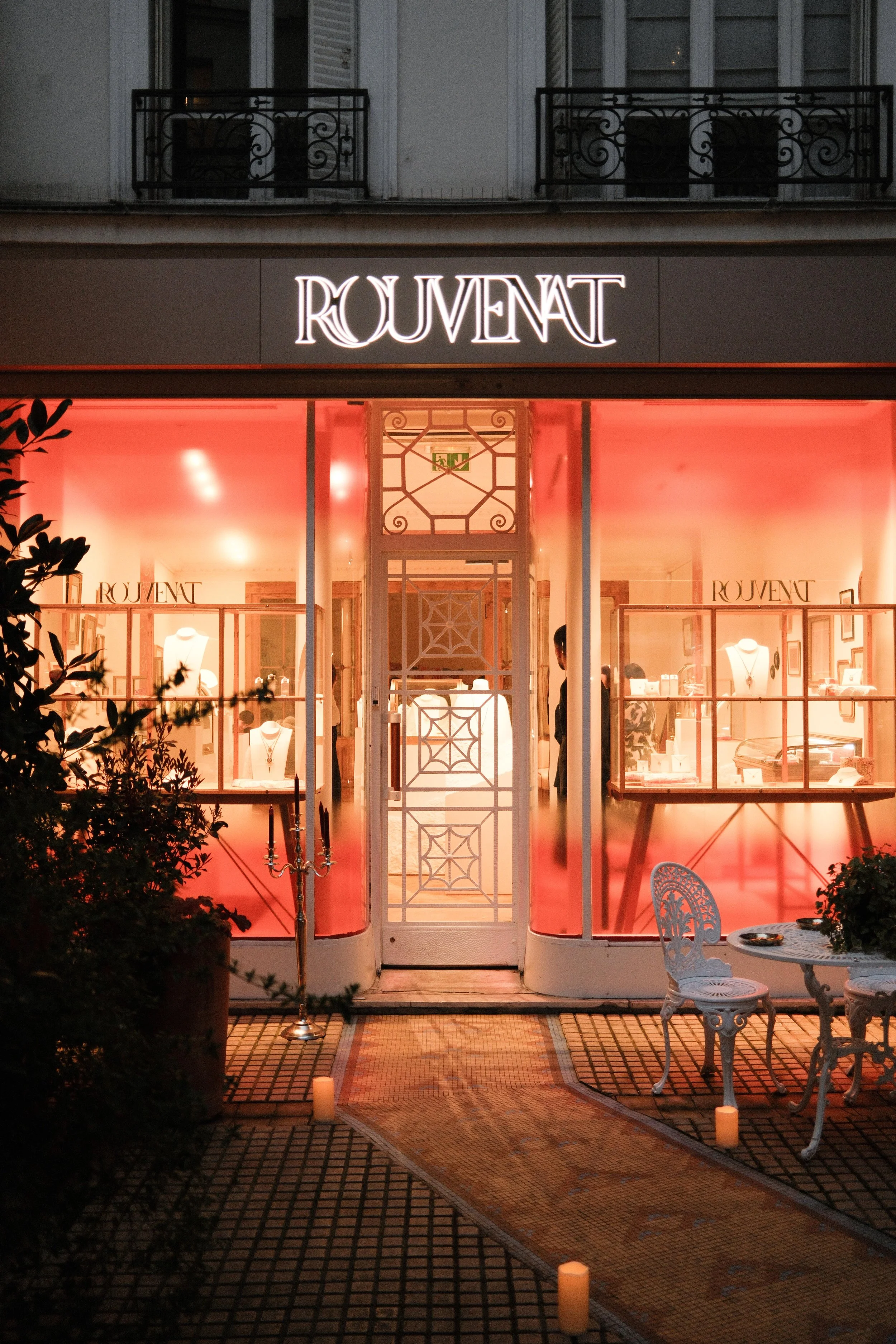  storefront with illuminated sign reading 'ROUENAT', pink lighting inside, outdoor white table and chairs, candles on the pavement outside.