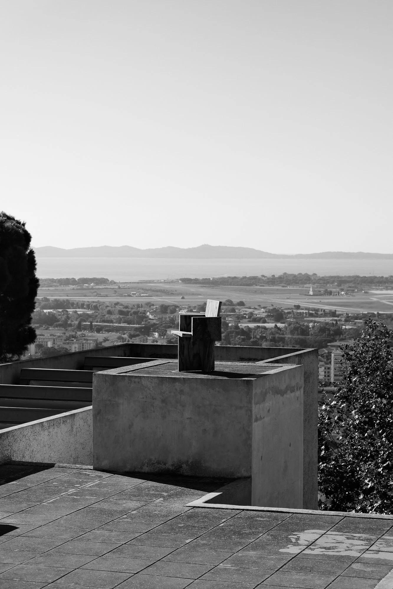 A black and white photo of a wooden chair on a rooftop with an expansive view of a cityscape, mountains in the distance, and a body of water on the horizon.