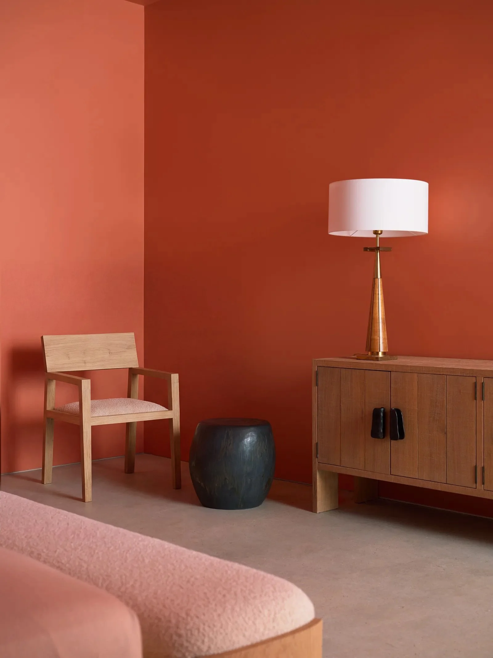 A room with a pink wall on the left and a darker brick red wall on the right. There is a wooden chair with a light wood frame and an upholstered pink seat in the foreground. A metallic black stool is partially visible behind a large dark vase, which is placed next to a wooden cabinet with closed doors and black handles. A tall, gold-colored table lamp with a white lampshade is on top of the cabinet.