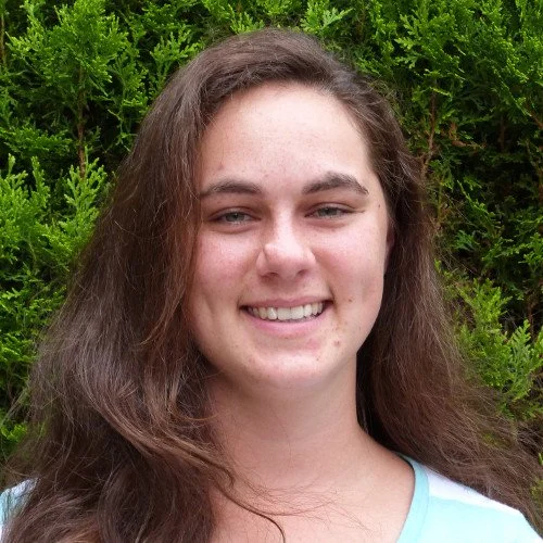 A young woman with long brown hair smiling outdoors in front of green foliage