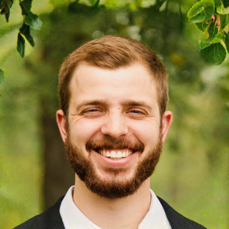 A smiling man with a beard and short brown hair outdoors, with green trees and foliage in the background.