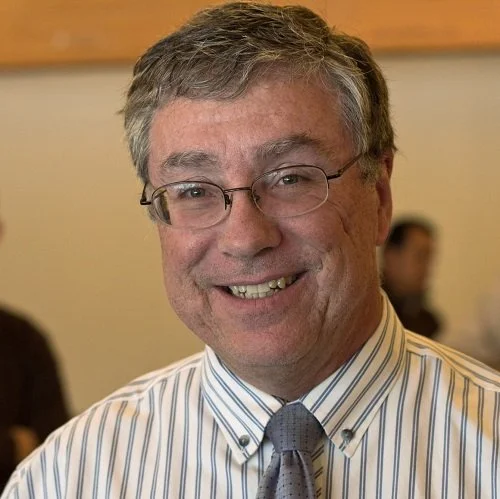 A smiling middle-aged man with glasses and gray hair, wearing a striped dress shirt and a tie.