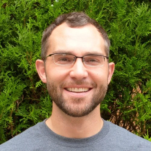 A smiling man with glasses and a beard standing outdoors in front of green bushes.