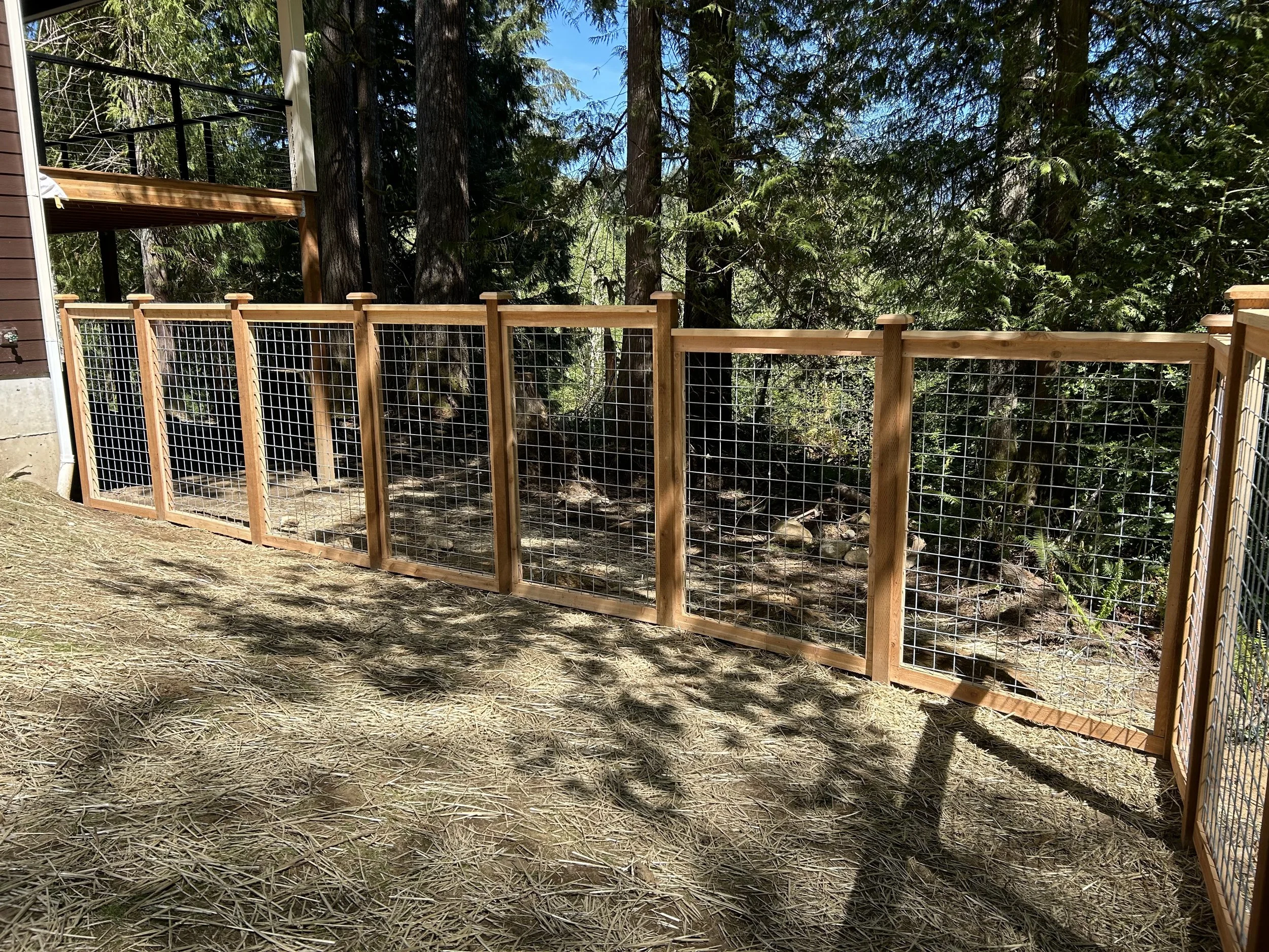 A wooden and wire mesh fence surrounds a wooded outdoor area next to a house with a brown exterior on a sunny day.