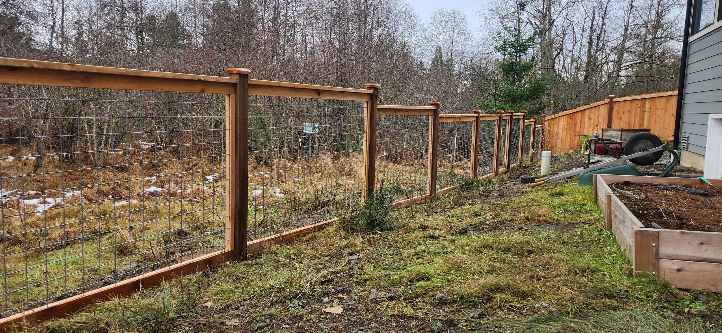 Newly constructed wooden and wire backyard fence along a grassy area, with a raised garden bed and gardening tools nearby.