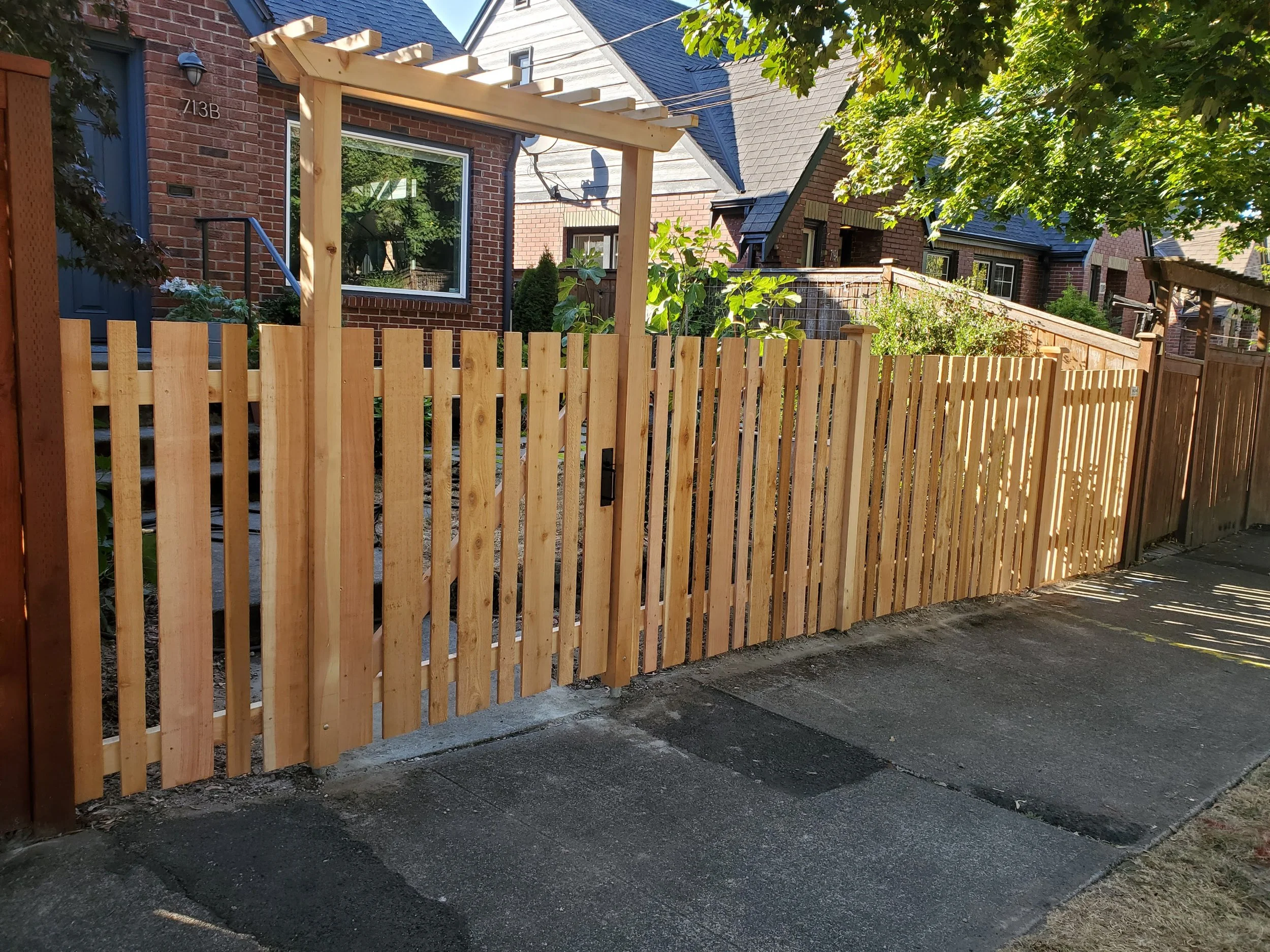 New wooden fence with a gate in front of a brick house with a porch and a large window, surrounded by trees and other houses in the background.
