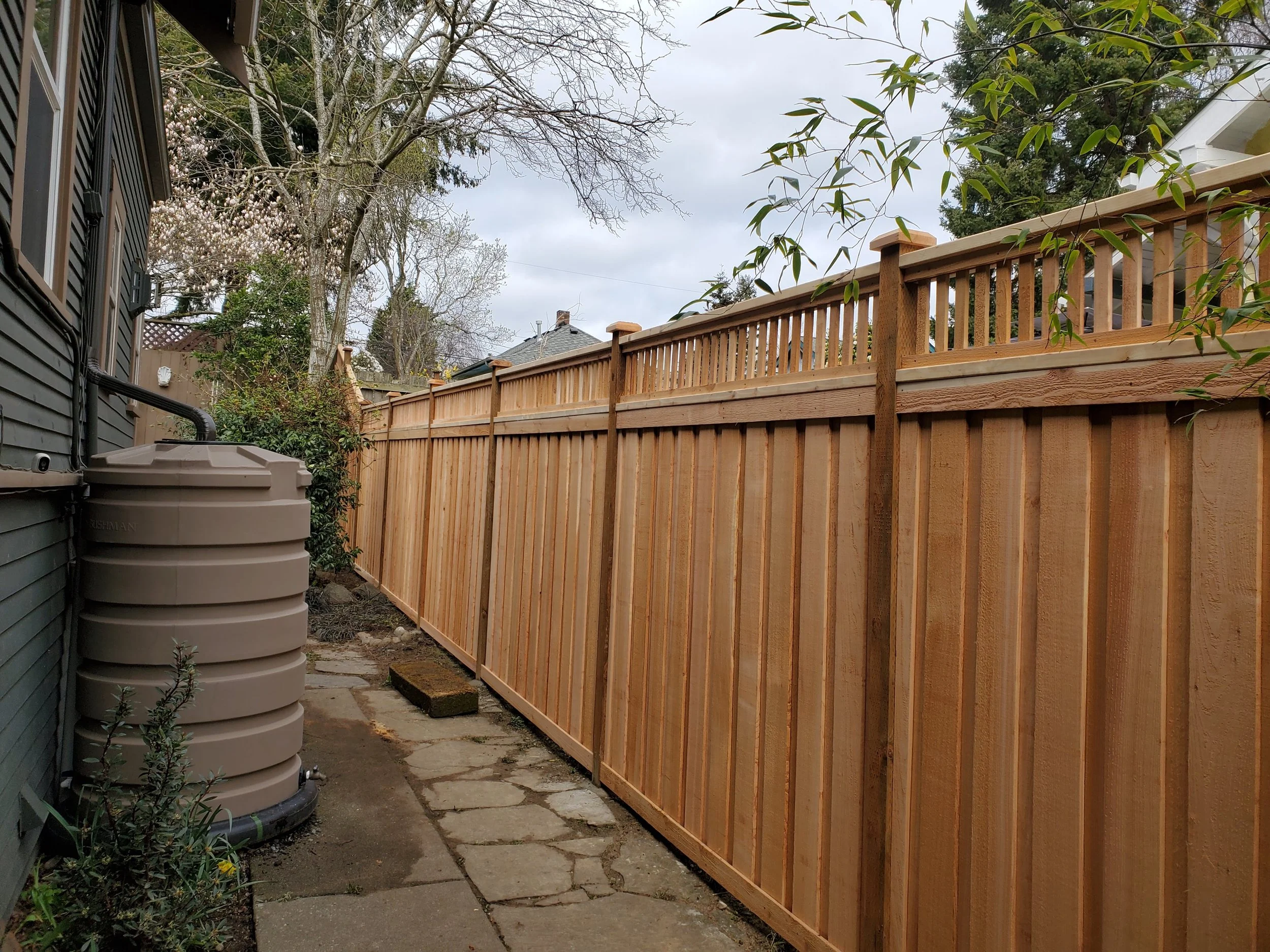 New wooden privacy fence along a narrow pathway beside a house, with trees and cloudy sky in the background.