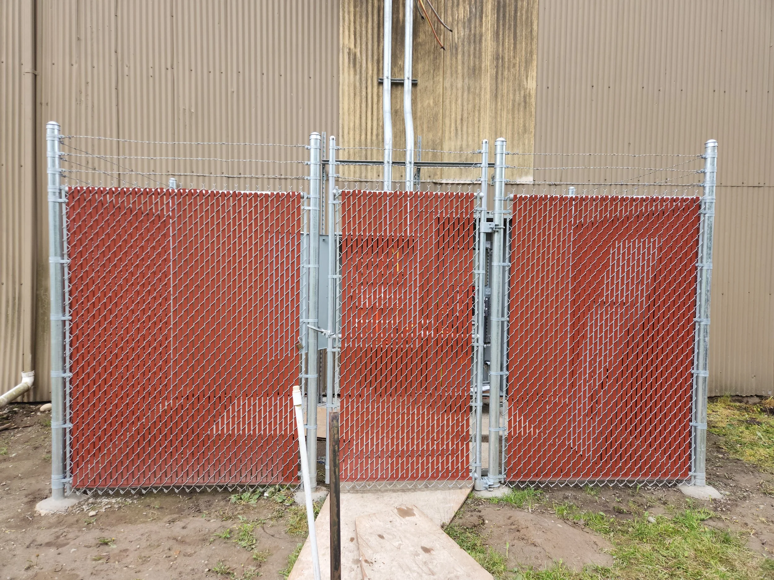 A fenced enclosure with three red panels and a small door in the middle, located outdoors against a corrugated metal wall.