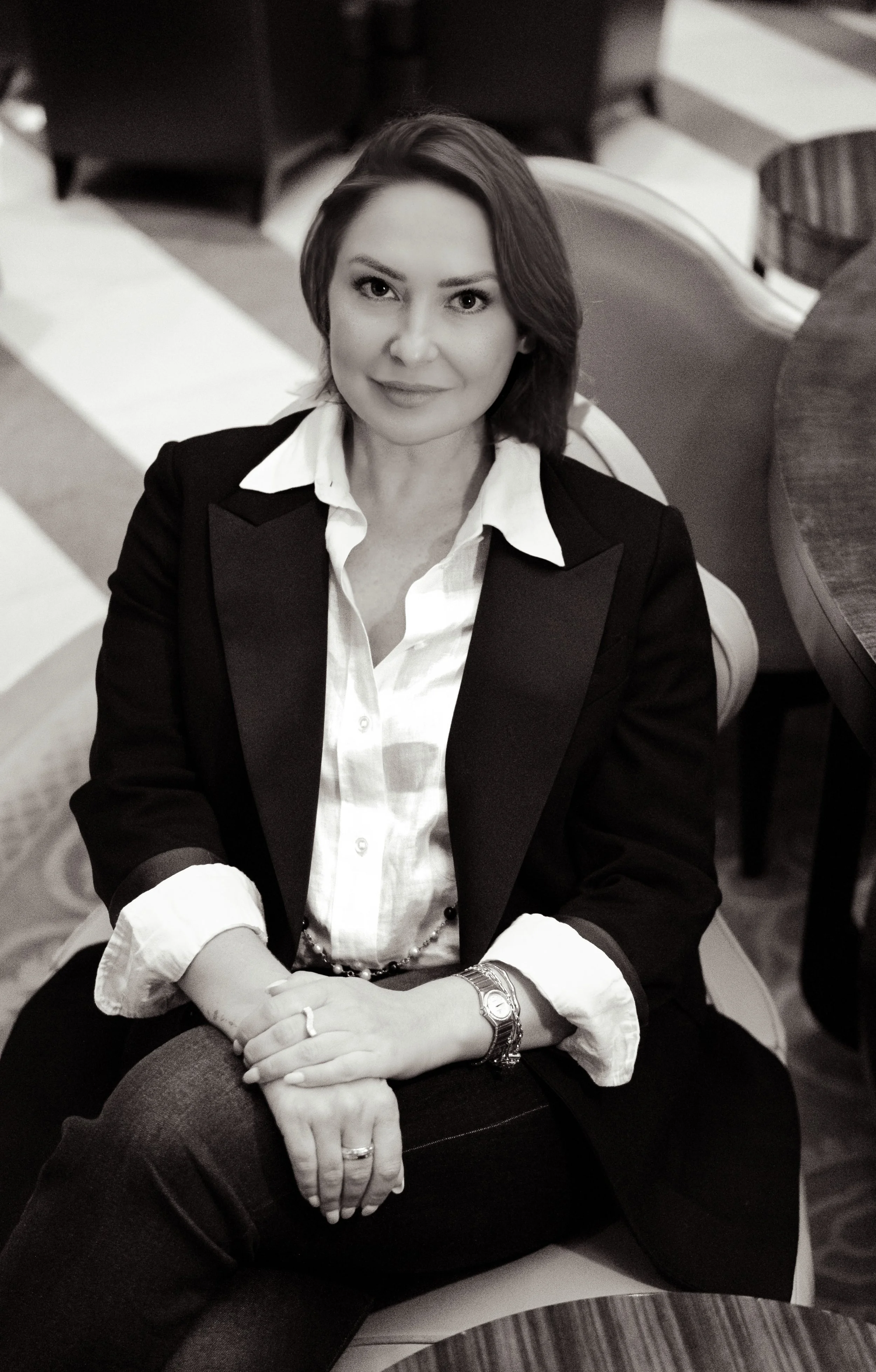 A woman with short hair wearing a tuxedo jacket over a white shirt, sitting at a table in a restaurant or hotel lobby.