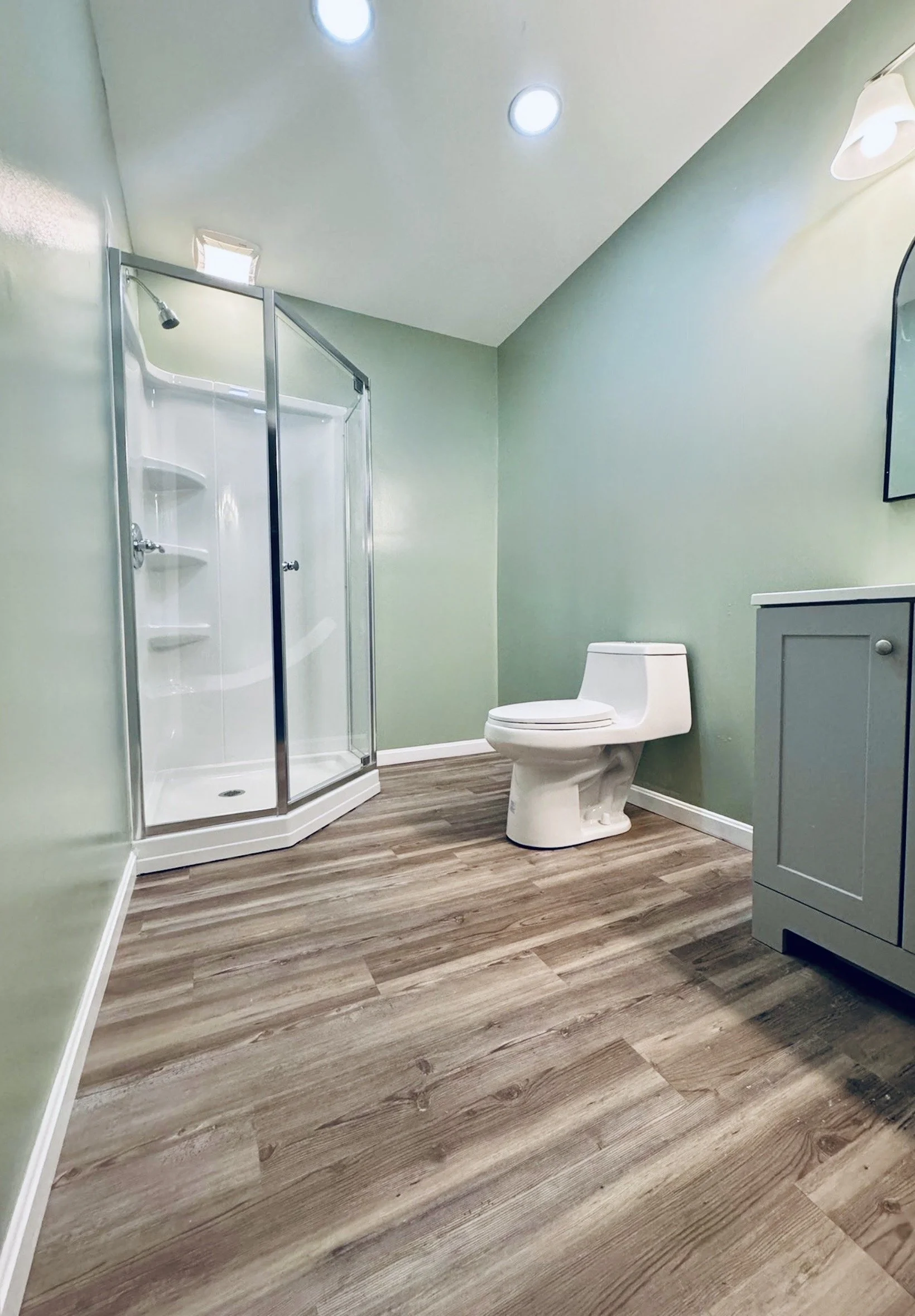A clean bathroom with a glass shower stall, a toilet, a small gray cabinet, and a mirror on the wall. The bathroom has wood-patterned flooring and light green walls.