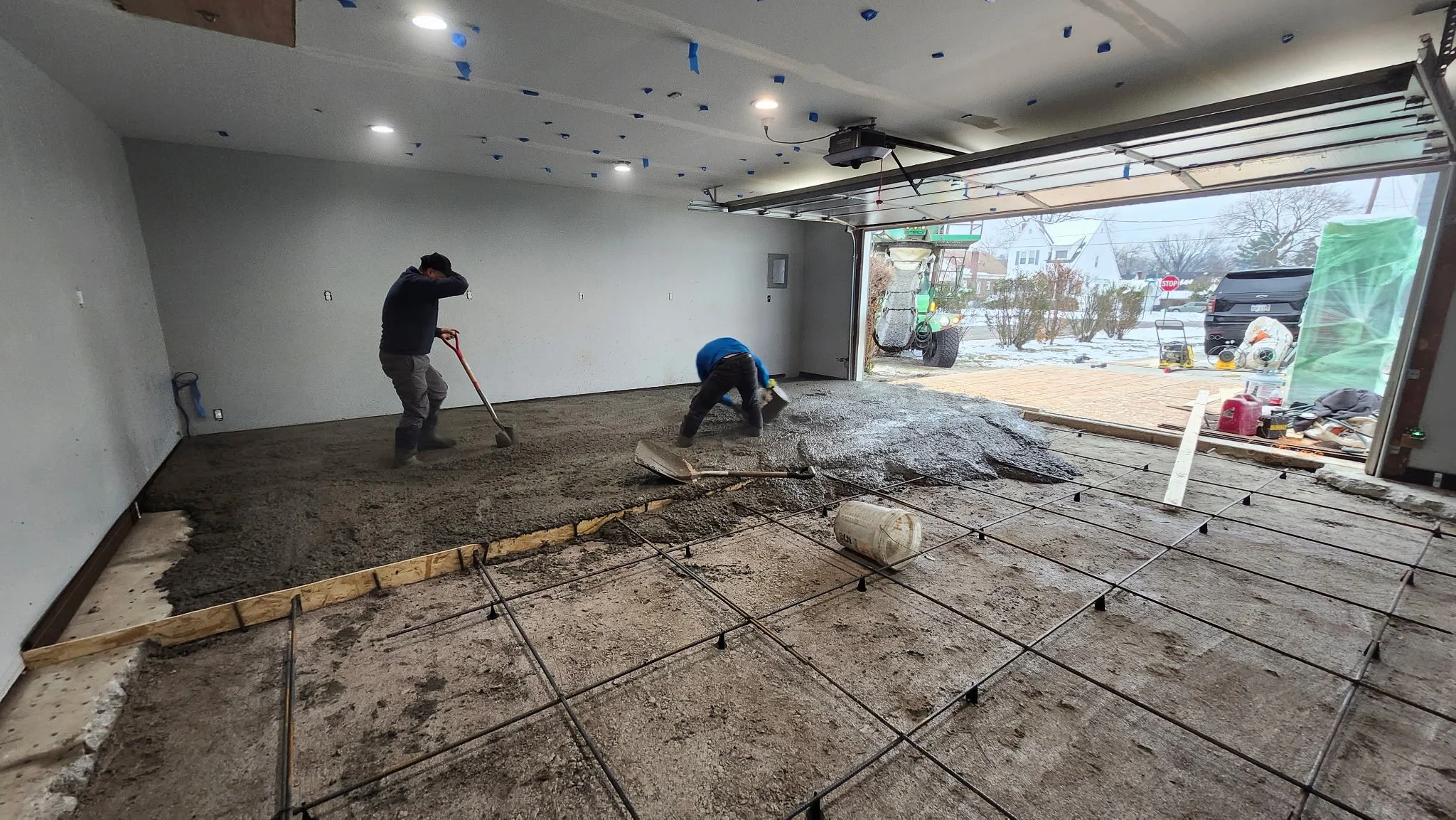 Two workers are leveling freshly poured concrete in a garage with an open door, revealing a snowy outdoor scene with vehicles and houses.