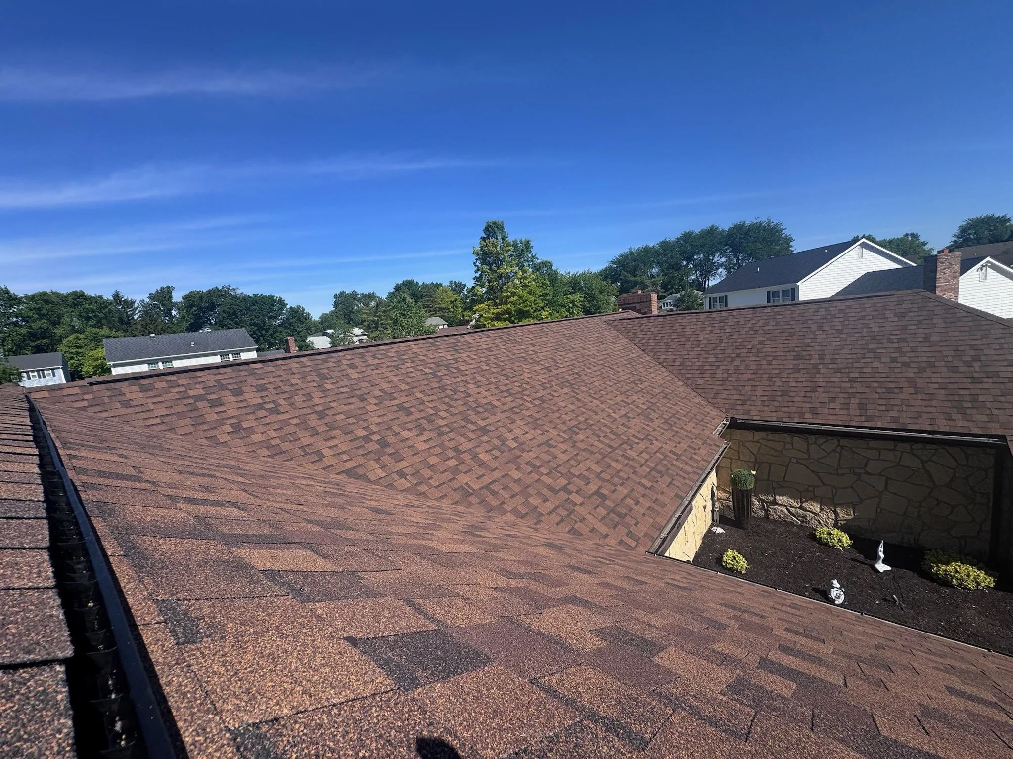 Overhead view of gray shingled roof of a house with trees and lawn in the background.