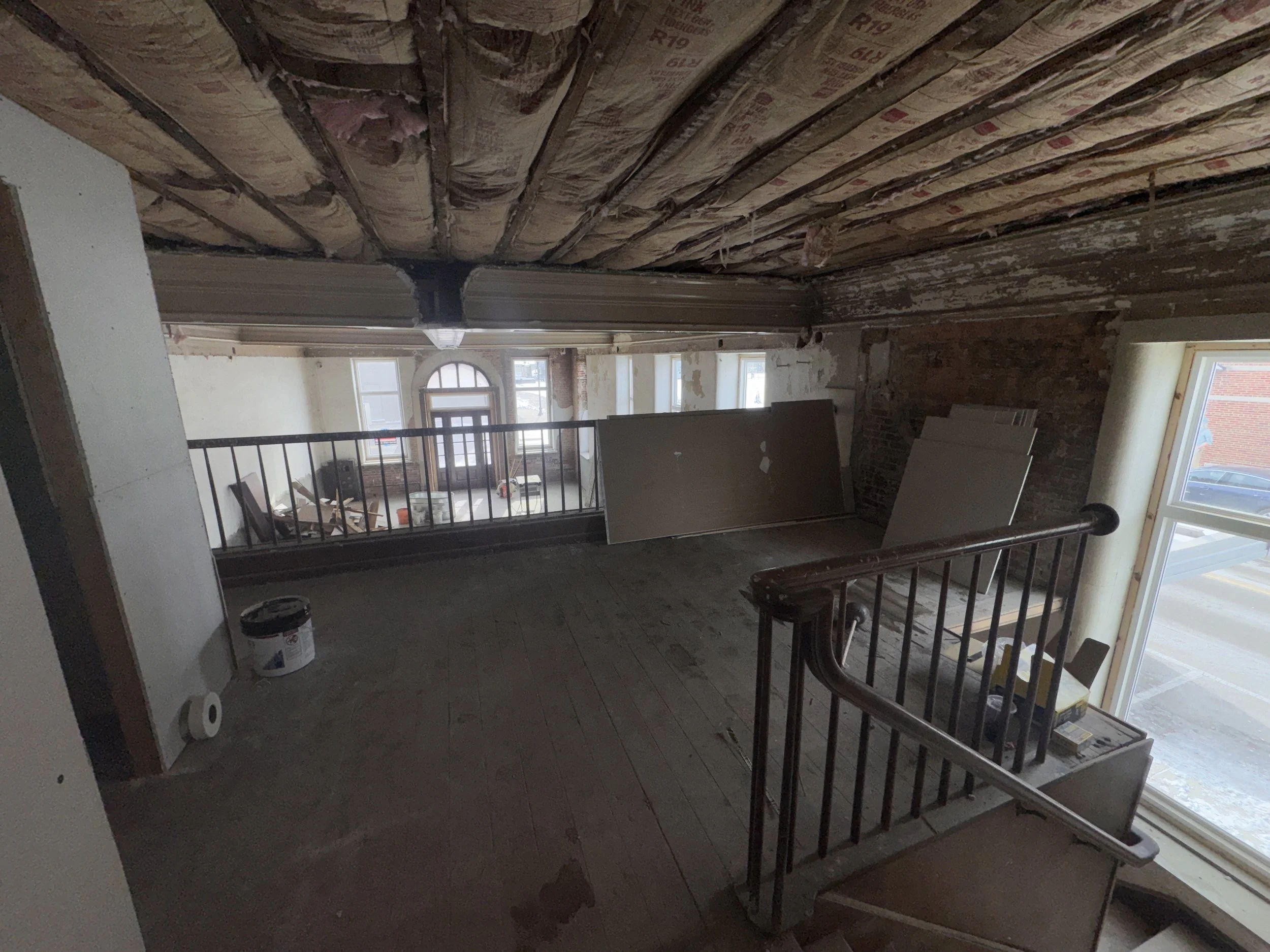Interior of a room under renovation, showing exposed wooden ceiling joists with insulation, a staircase with a wooden handrail, large windows, and construction materials and tools around.