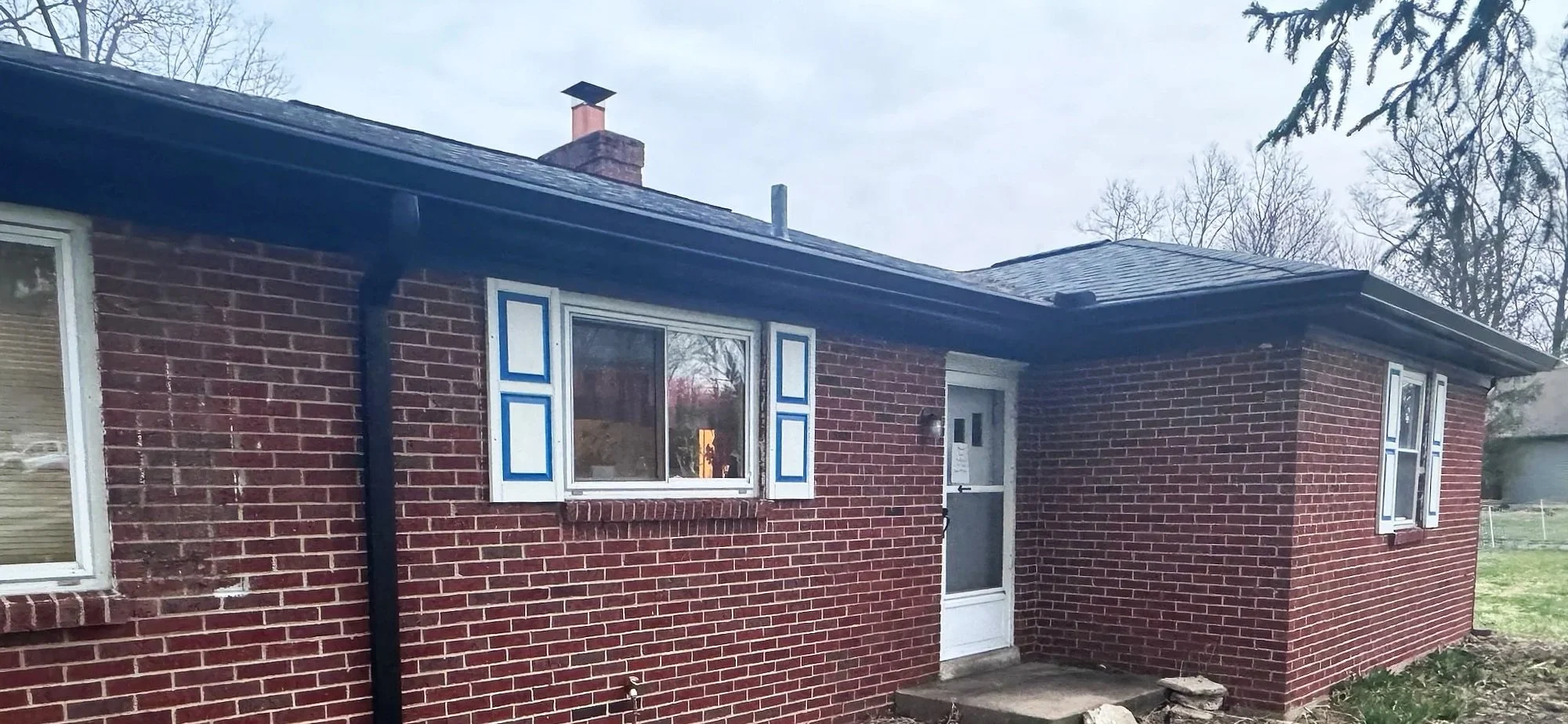 A brick house with three windows, blue and white shutters, a white door with a small window, and a black gutter, with trees and grass in the yard.