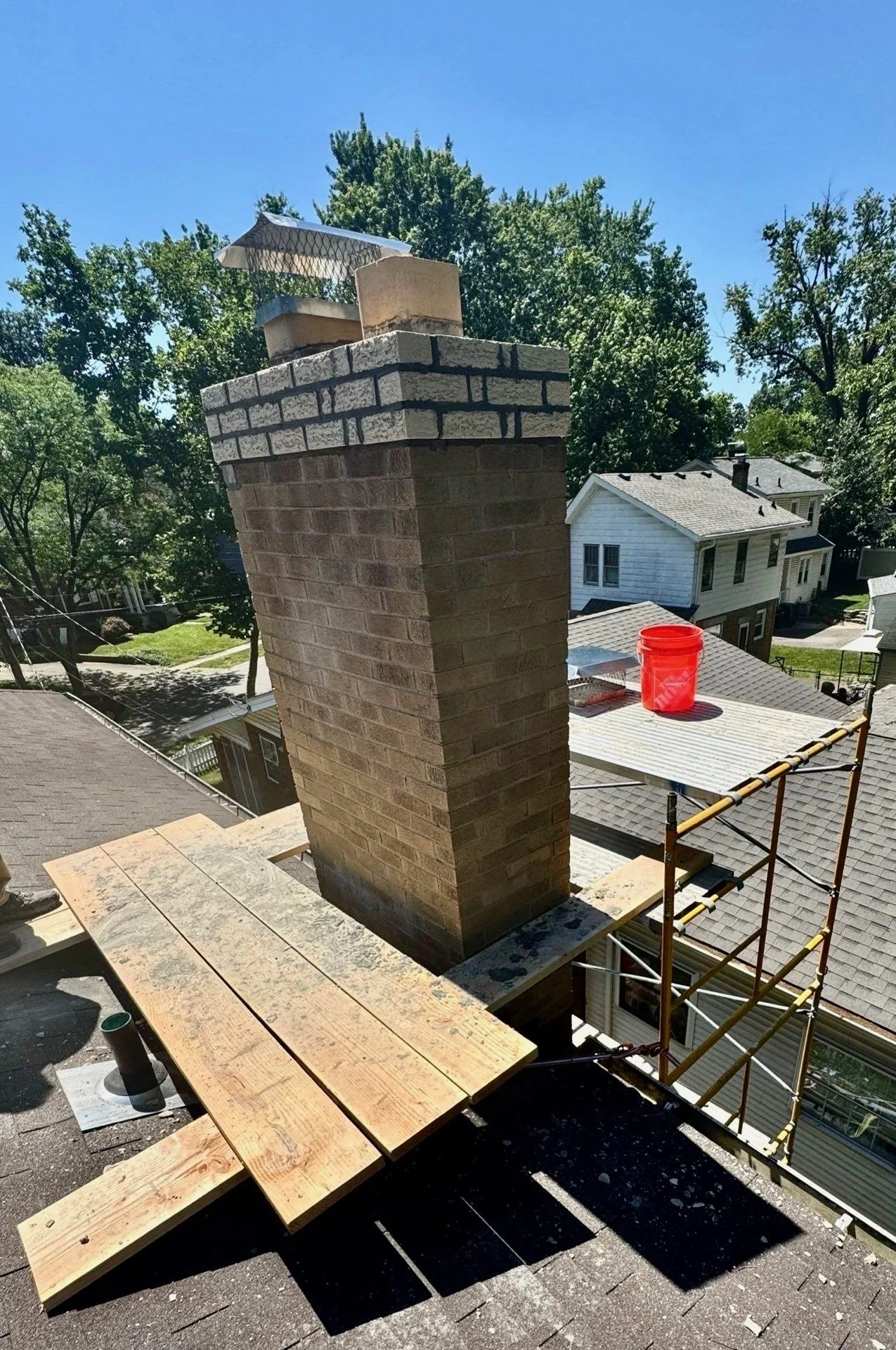 View of a brick chimney on a rooftop with construction scaffolding and wooden planks nearby, in a residential neighborhood with trees and houses under a blue sky.
