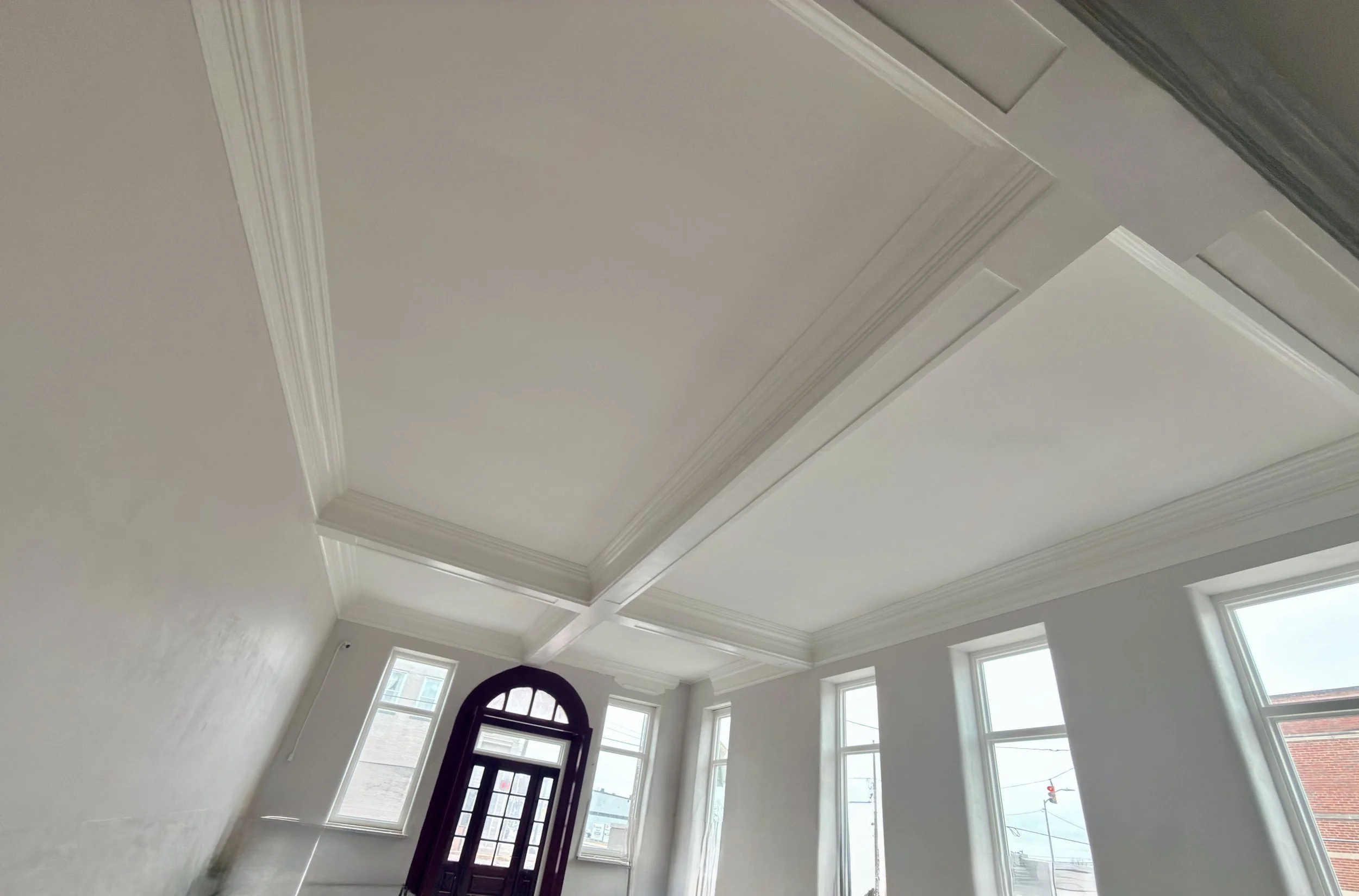 Interior view of a room with large windows, a decorative ceiling, and a front door with glass panels, during daytime.