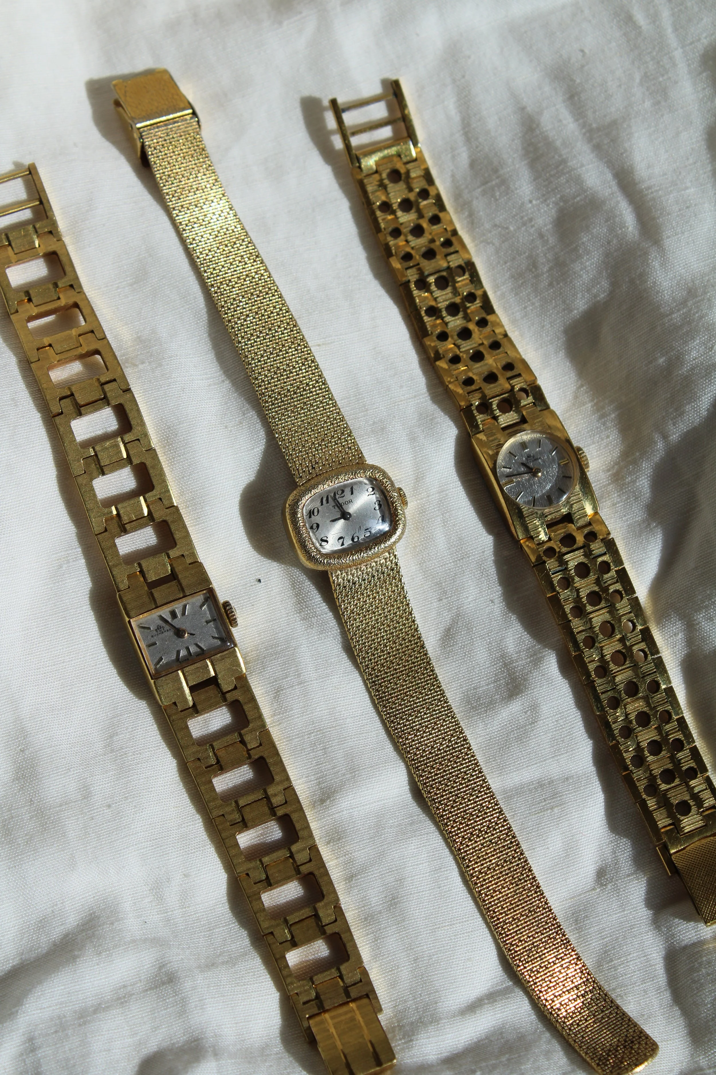 Three gold wrist watches with metal bands laid on a white fabric background.