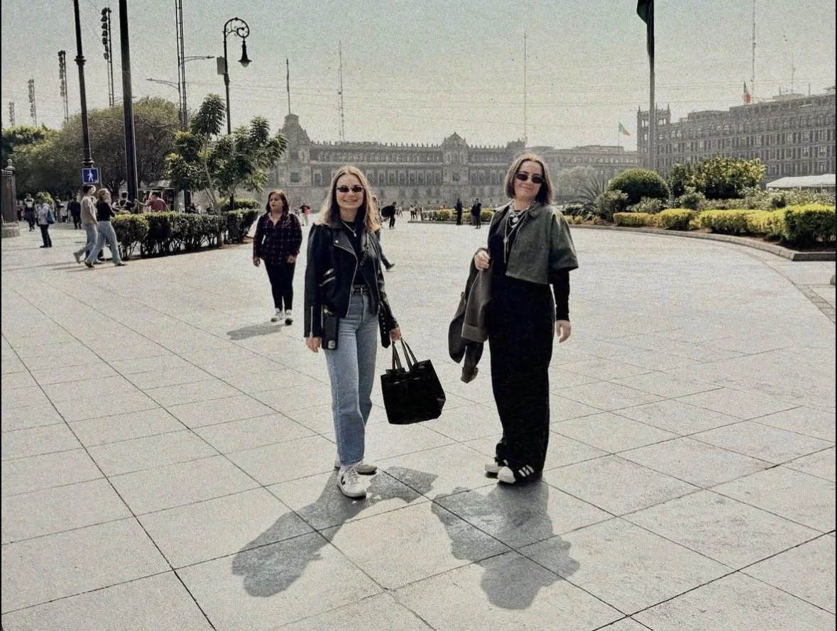 Two women smiling and walking in an open plaza area with large building in the background, trees, and lampposts.